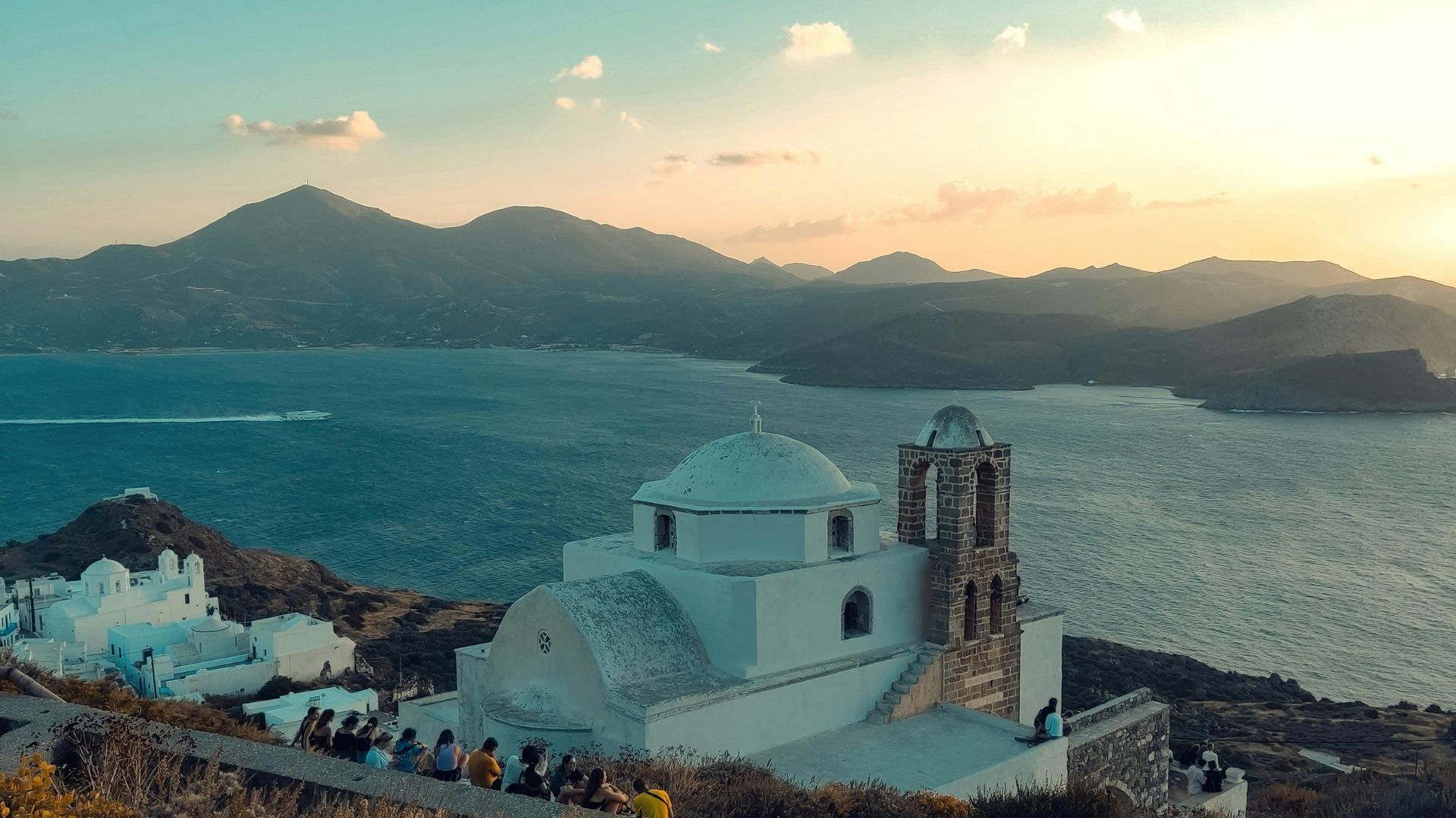 A picturesque white church with a brick bell tower sits atop a hill overlooking the Aegean Sea in Milos, Greece.