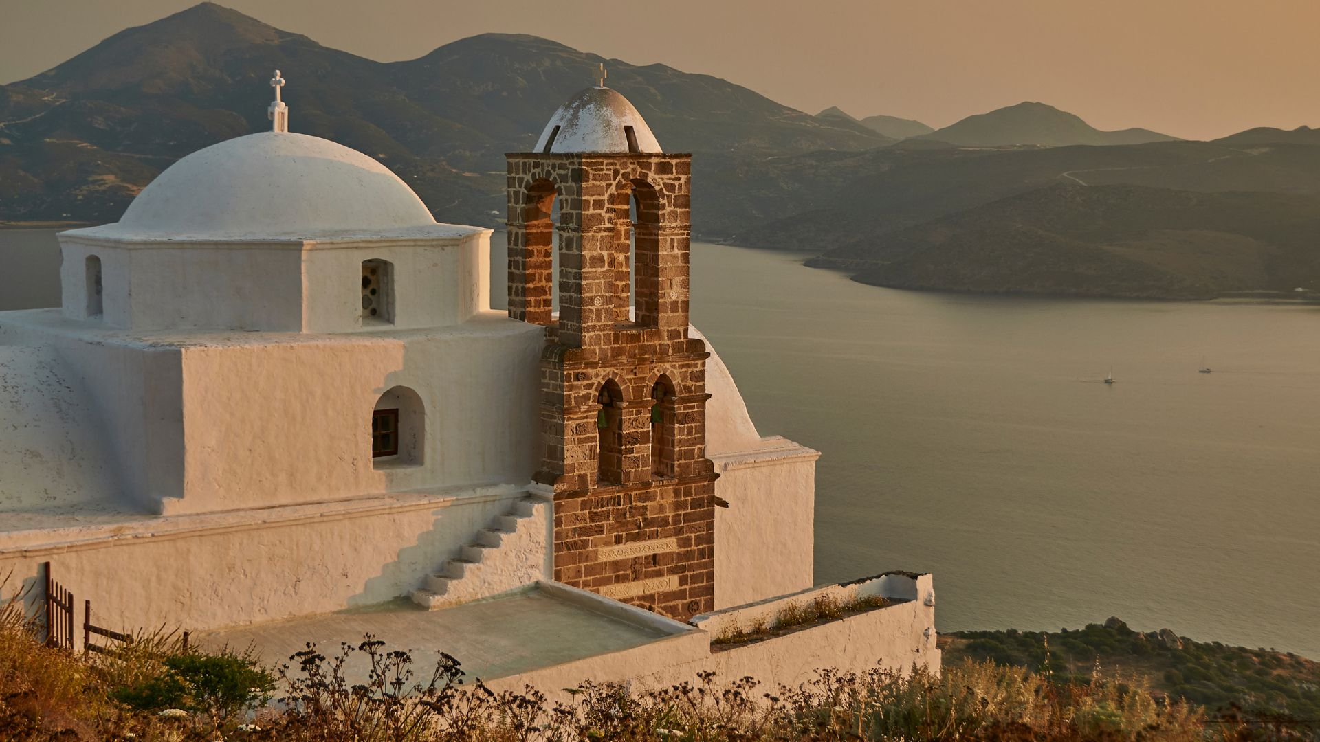 A picturesque white church with a brick bell tower sits atop a hill overlooking the Aegean Sea in Milos, Greece.