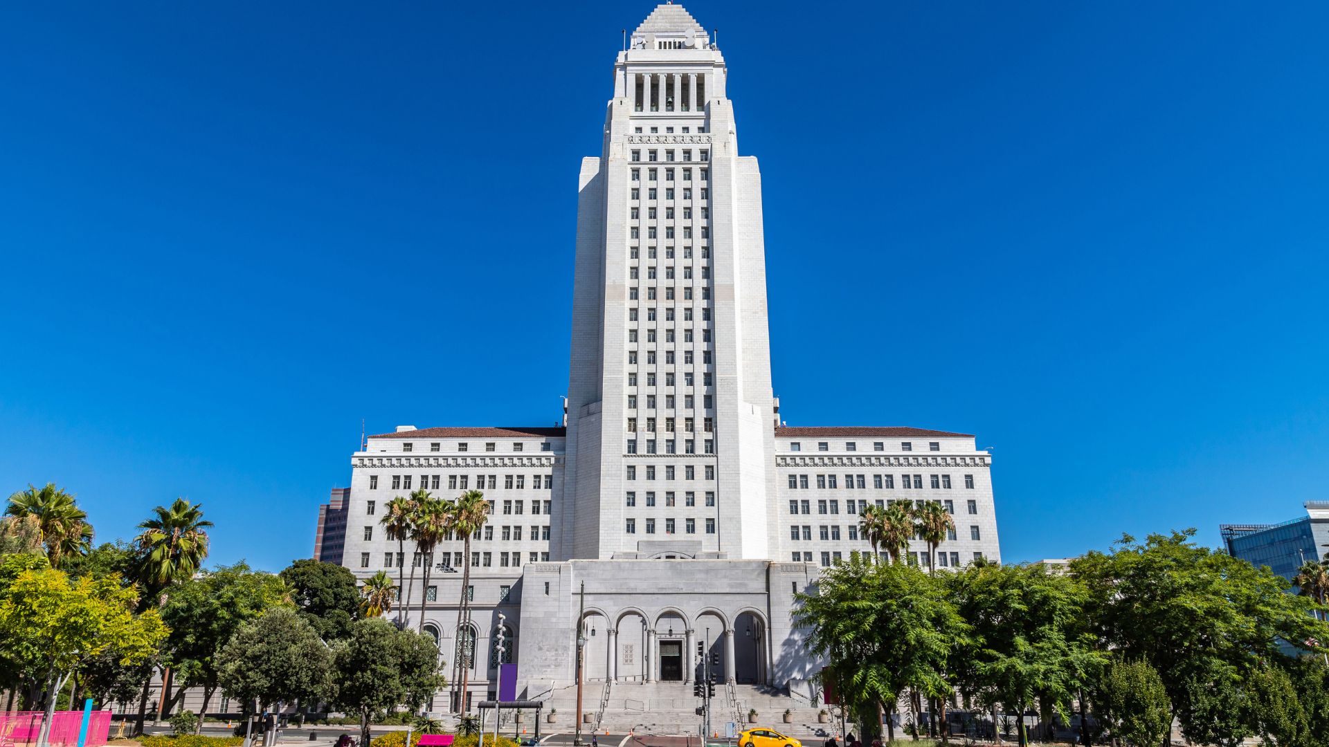 Los Angeles City Hall, a historic 32-story Art Deco building in downtown Los Angeles, California, under a clear blue sky, surrounded by lush greenery and palm trees.