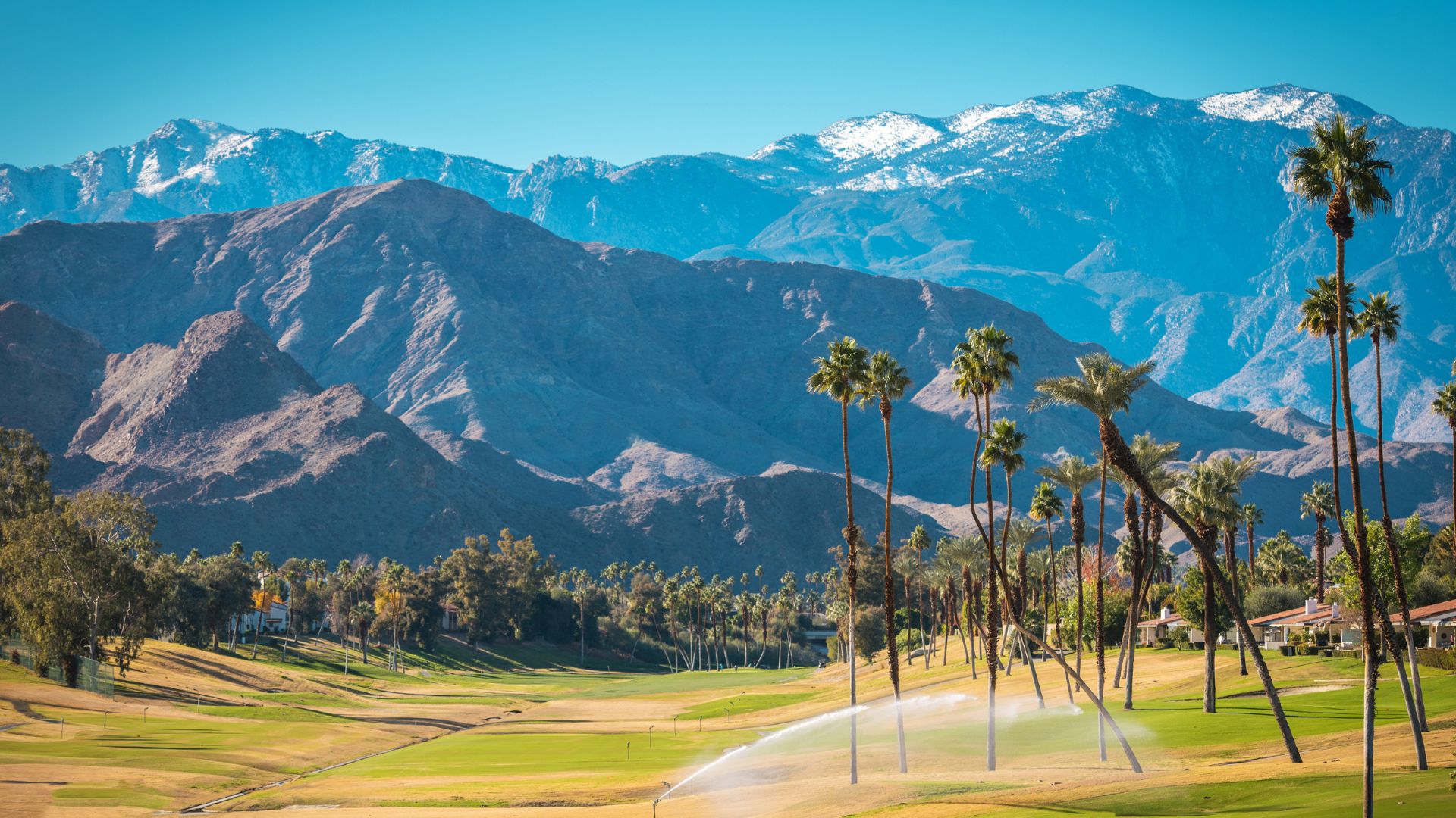A lush green golf course with palm trees in the foreground, set against a backdrop of towering, snow-capped mountains under a clear blue sky. Sprinklers are visible watering the course.