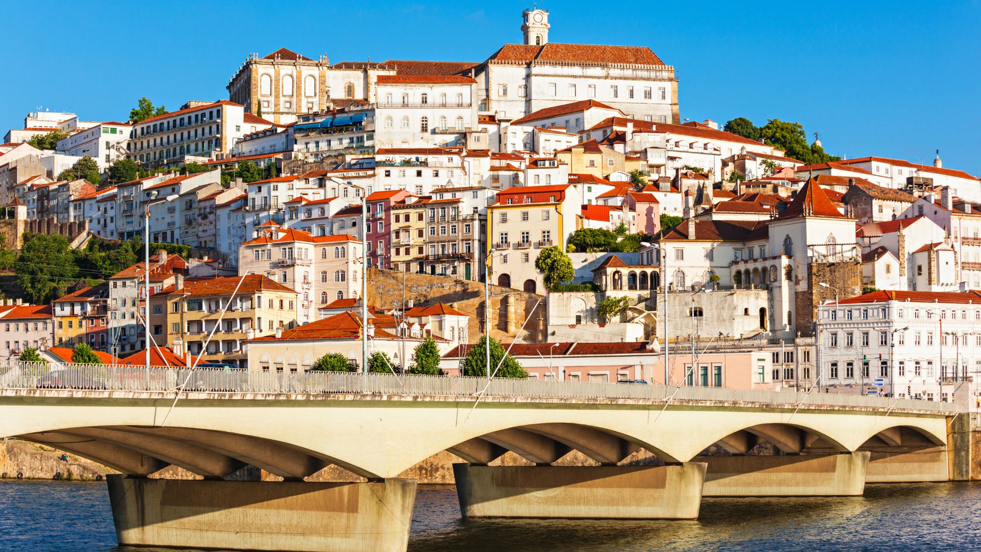 A panoramic view of Coimbra, Portugal, featuring a bridge over the Mondego River in the foreground and the historic city with white buildings and a prominent university complex on a hill in the background under a clear blue sky. 