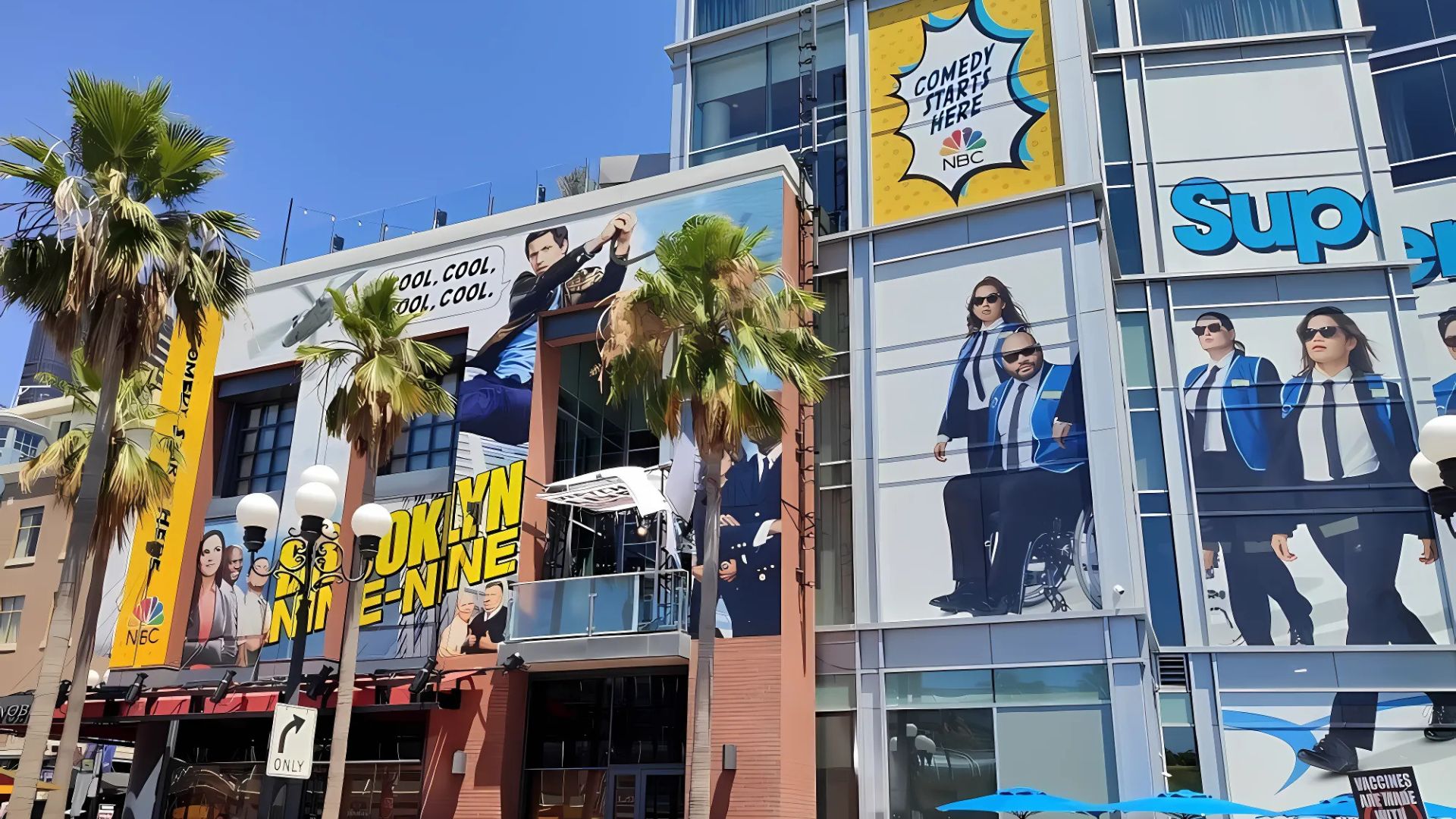 A vibrant street scene in San Diego during a large event, featuring buildings adorned with massive banners promoting NBC shows like "Brooklyn Nine-Nine" and "Superstore,"