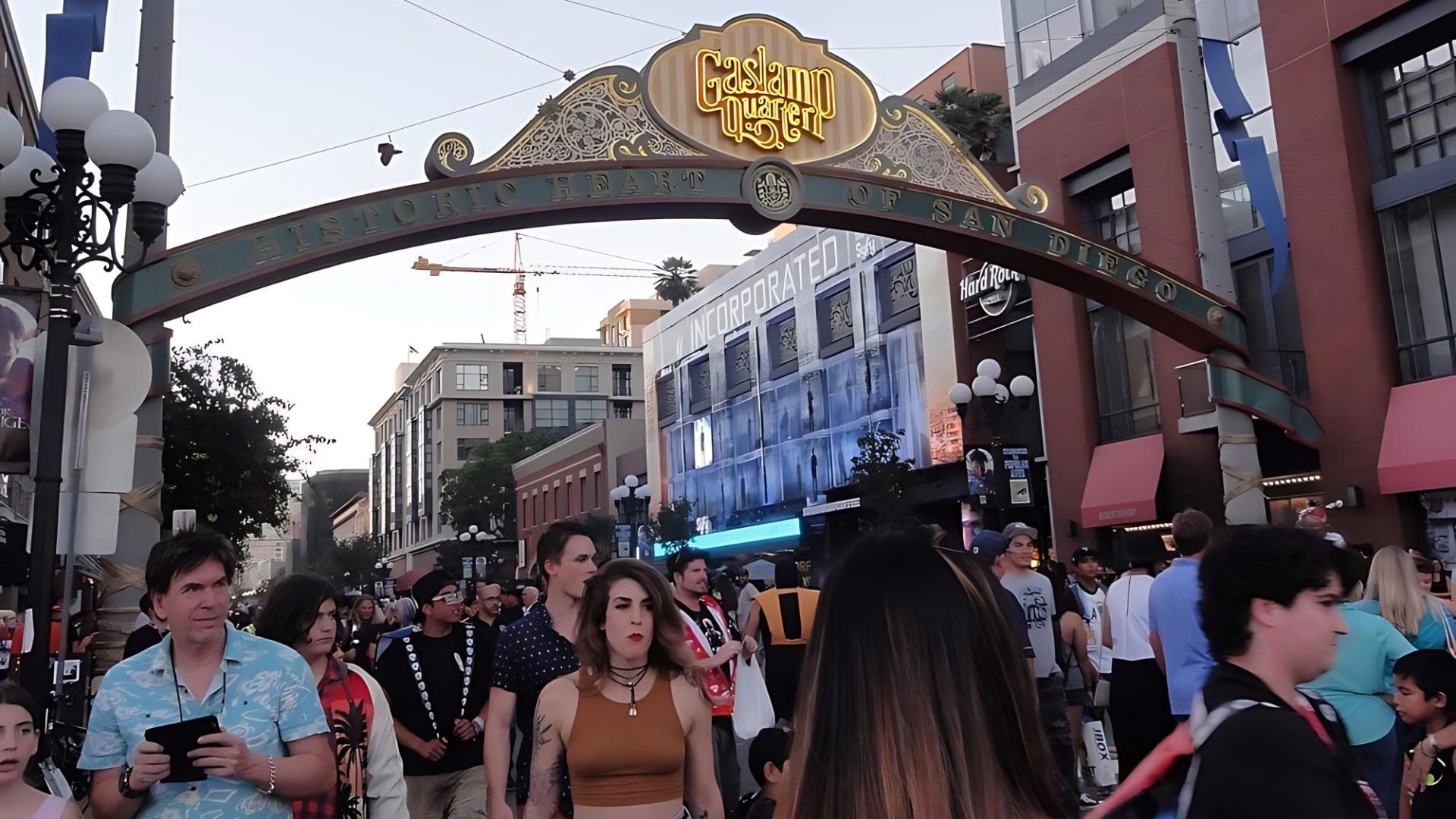 A crowded street scene in San Diego's Gaslamp Quarter, featuring the iconic Gaslamp Quarter archway with "Historic Heart of San Diego" inscribed, and various buildings in the background, including a Hard Rock Hotel and a building labeled "INCORPORATED."