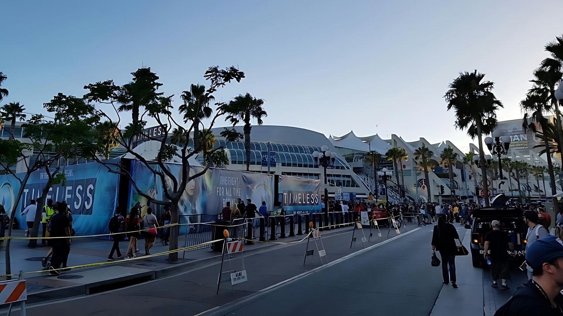 A wide shot of the San Diego Convention Center during Comic-Con, showing crowds of people, banners, palm trees, and a street with vehicles under a clear sky.