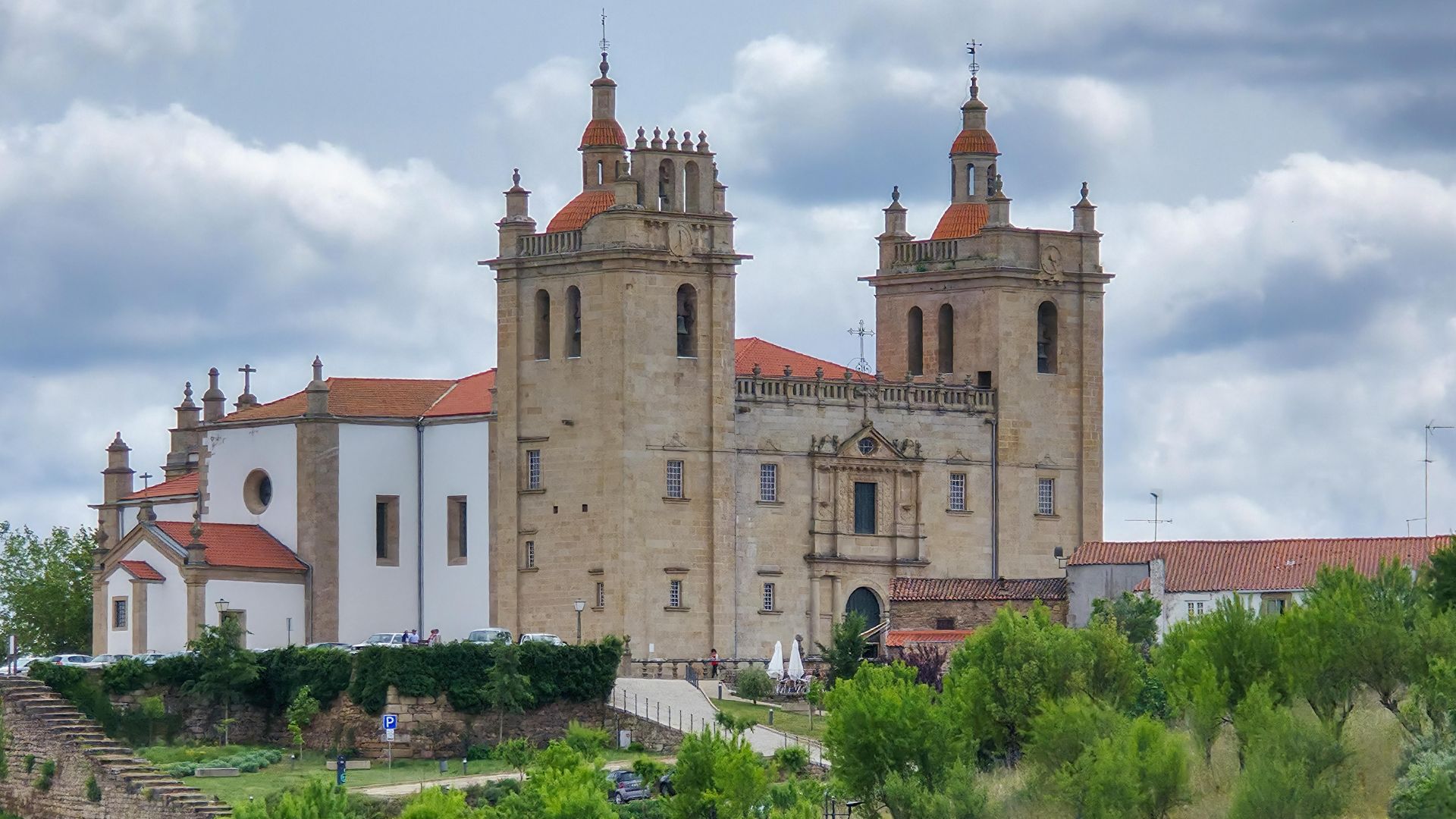 A large, historic stone cathedral with two prominent towers topped with red roofs, situated on a hill with green foliage in the foreground under a cloudy sky.