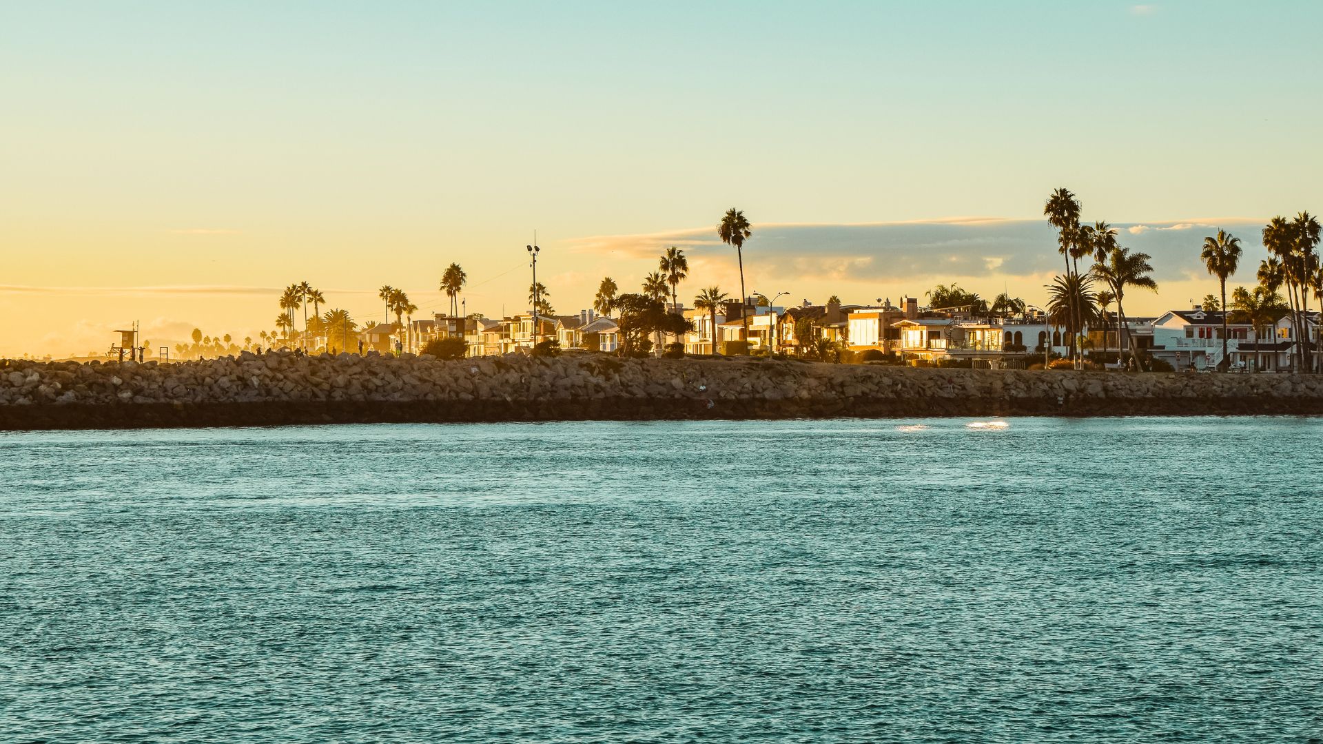 A wide shot of Newport Beach, California, at sunset, showing the ocean with gentle waves and two people on surfboards in the foreground. In the background, the sandy beach is visible with several people walking and buildings with palm trees lining the coastline. 