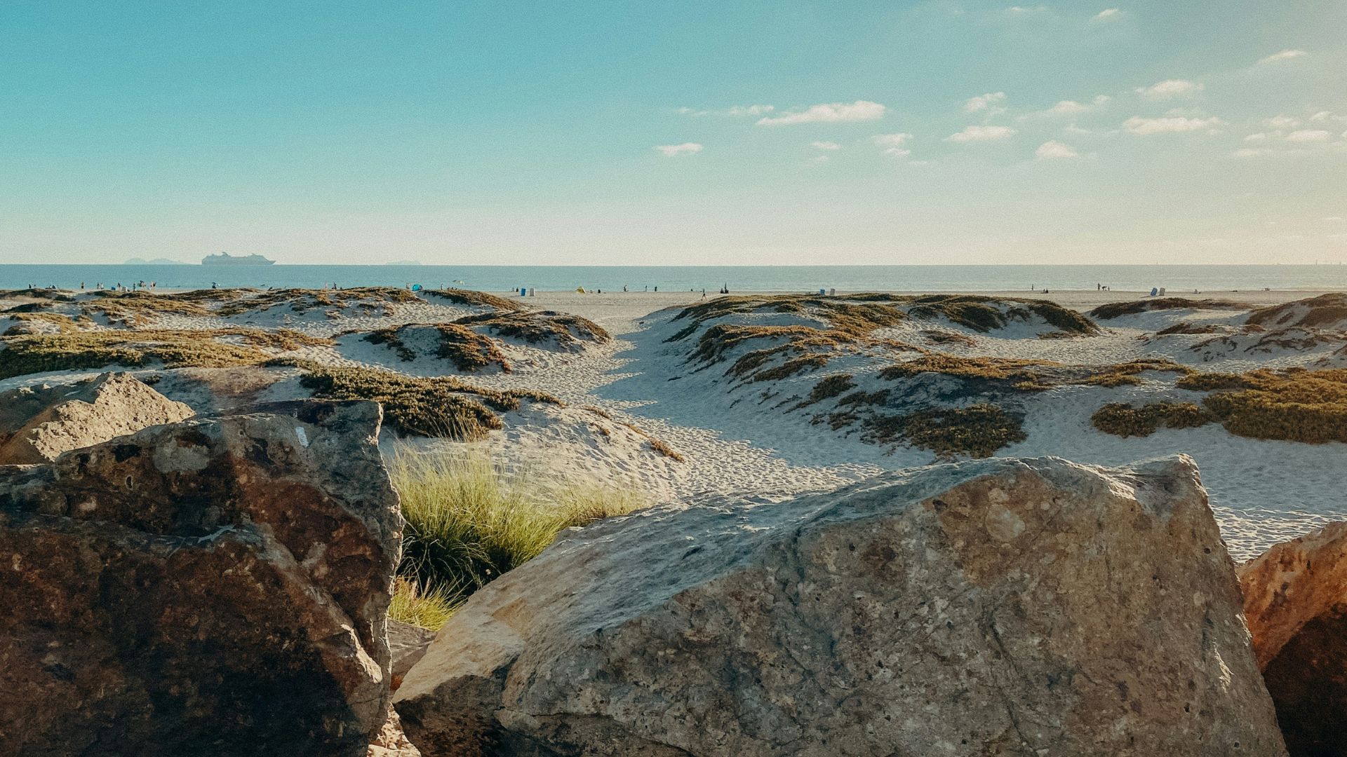 A wide-angle shot of a sandy beach with large rocks in the foreground, leading to gentle dunes covered with sparse vegetation, and the ocean with people in the distance under a bright sky. 