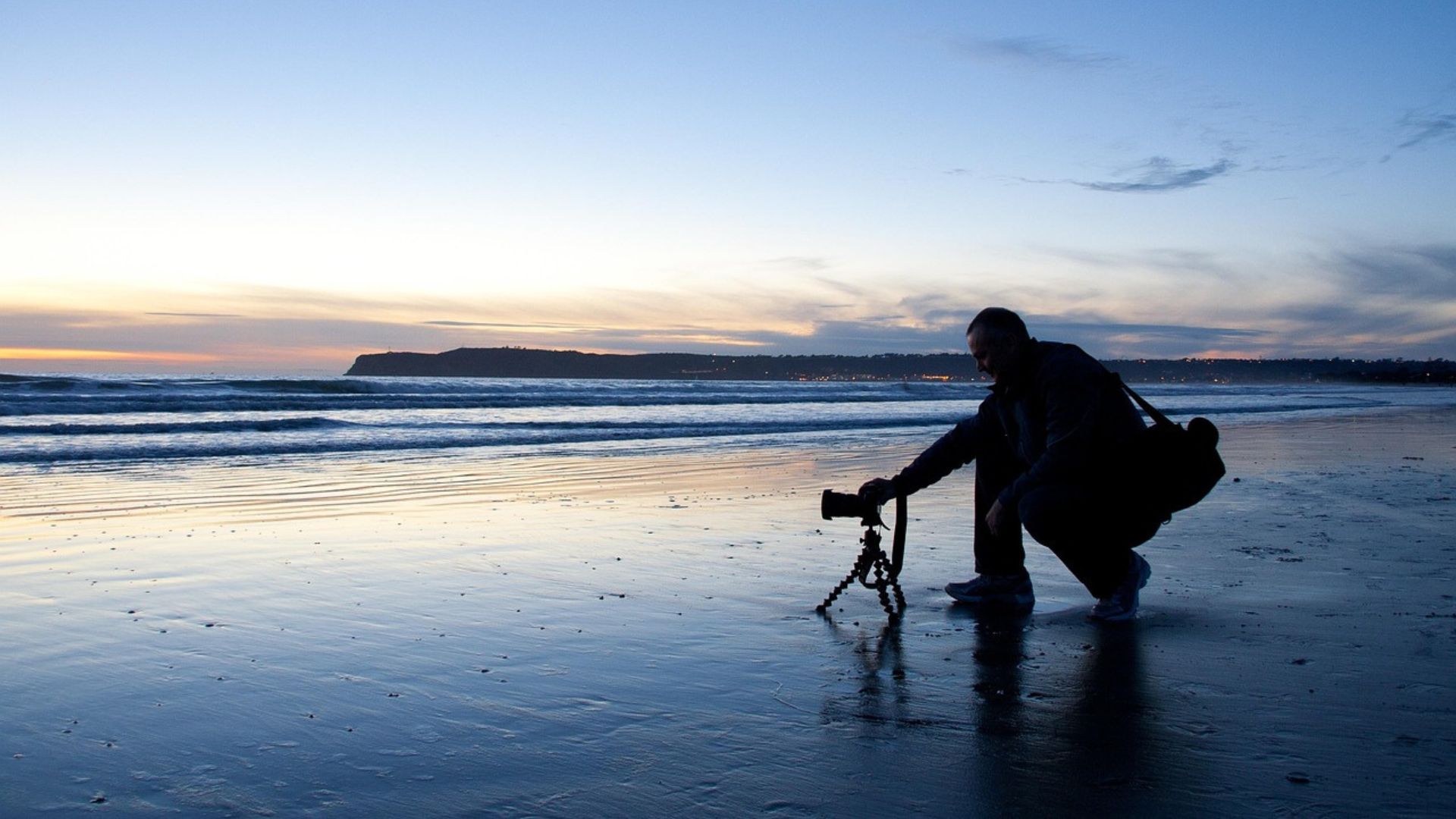 A silhouette of a photographer crouches on a wet beach at dusk, adjusting a camera on a small tripod, with waves gently rolling in and the distant lights of a city and landmass under a colorful sky.