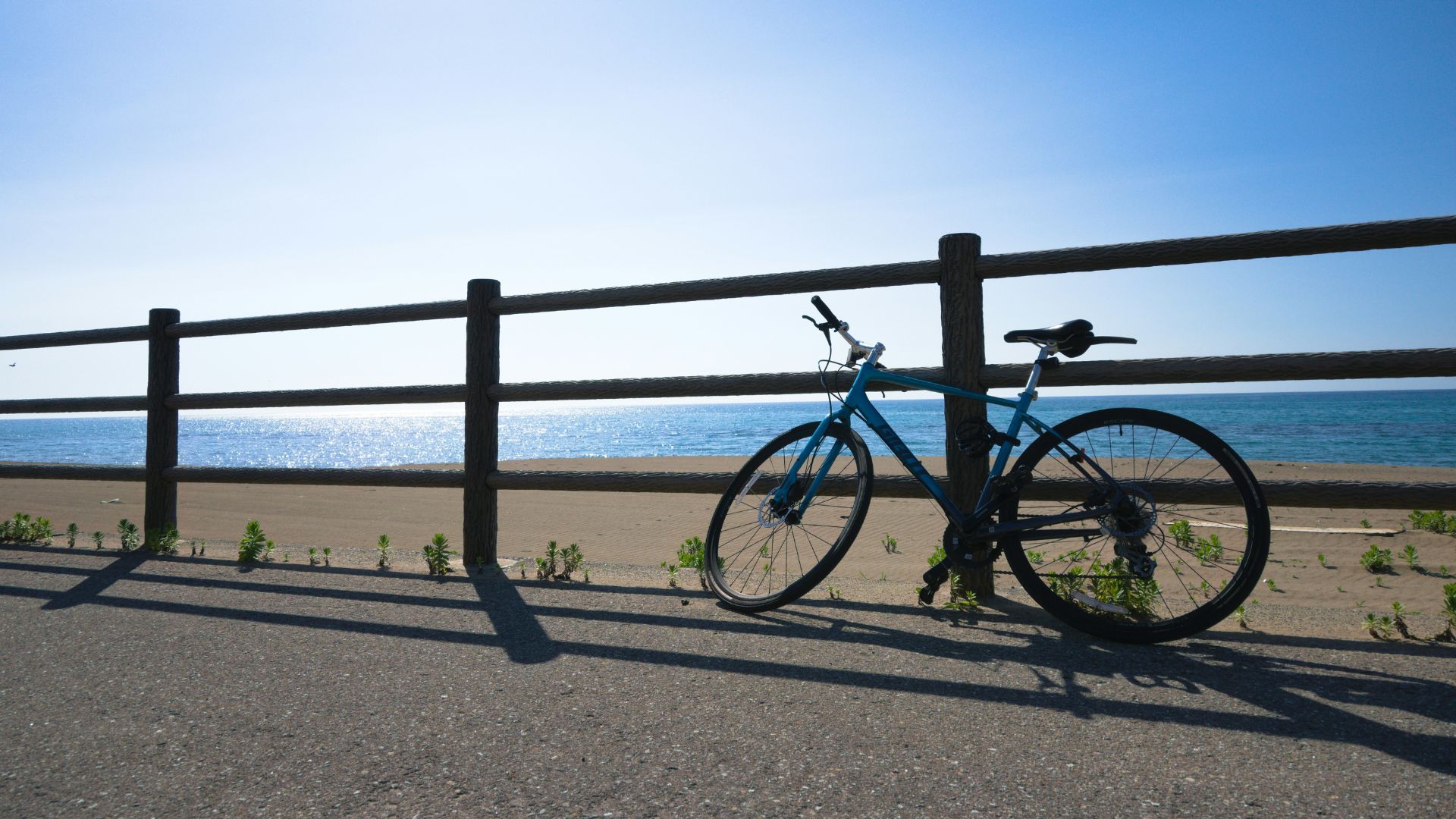 A blue bicycle leans against a wooden fence alongside a paved path, with a sandy beach and the ocean visible in the background under a bright, sunny sky.