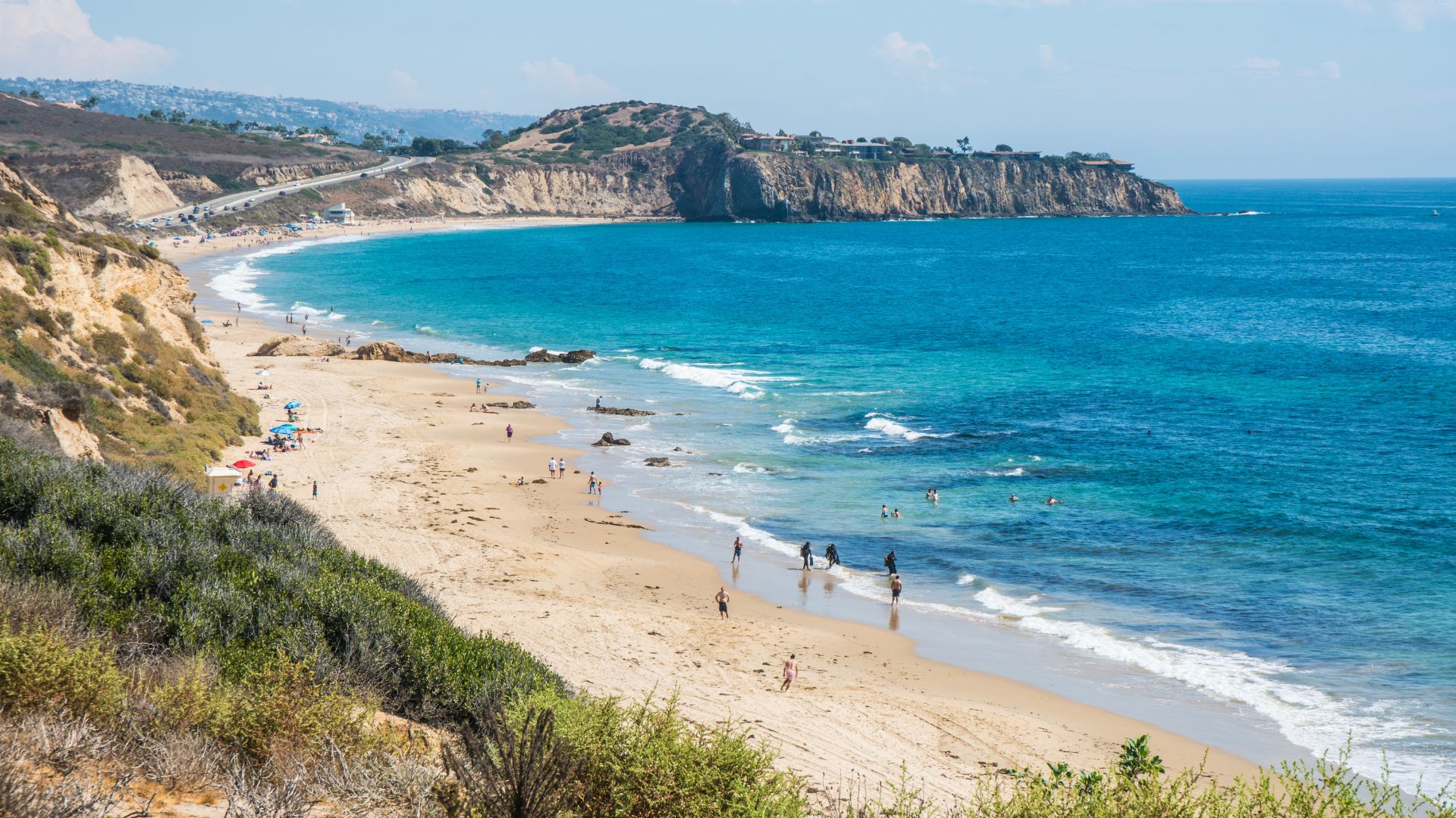 A wide-angle view of Crystal Cove State Park beach in Orange County, California, showing a long stretch of sandy beach with gentle waves, a clear blue ocean, and a rugged coastline with cliffs and lush green vegetation under a bright, sunny sky. Several people are visible along the shoreline.