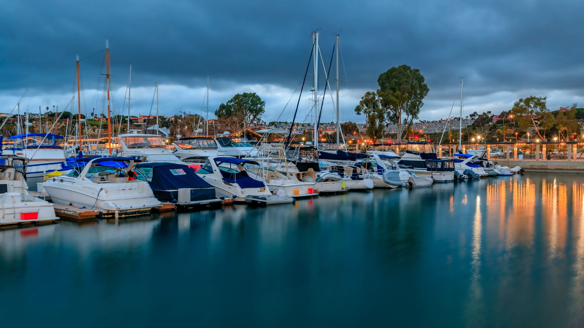 A nighttime scene at Dana Point Harbor, featuring numerous boats and yachts docked in the calm water under a dark, cloudy sky. Lights from the harbor and distant buildings reflect on the water's surface.