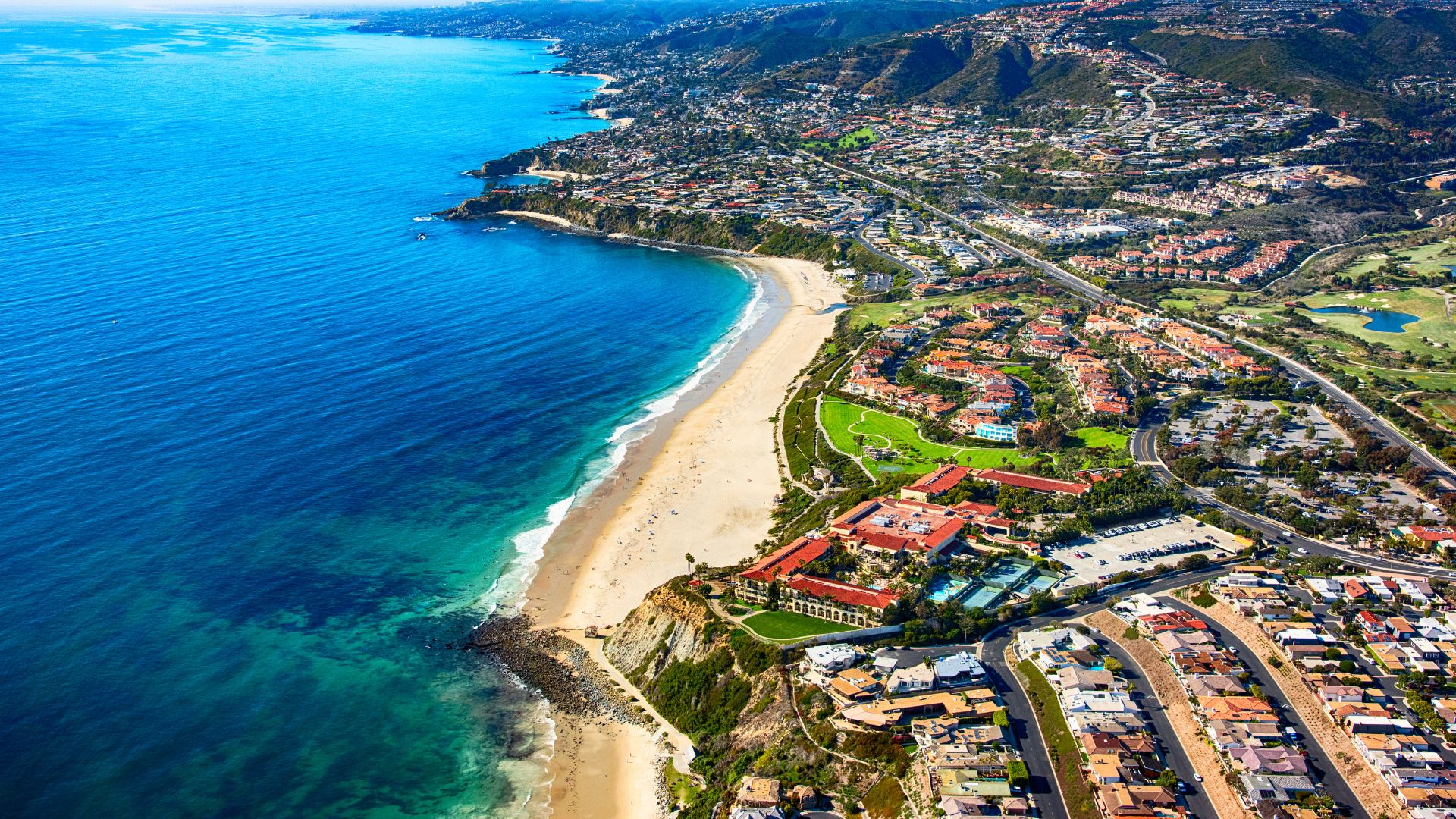 An aerial view showcases a vibrant coastal landscape featuring a wide, sandy beach bordering a clear blue ocean, with a developed shoreline containing residential areas, roads, and greenery extending towards rolling hills in the background.