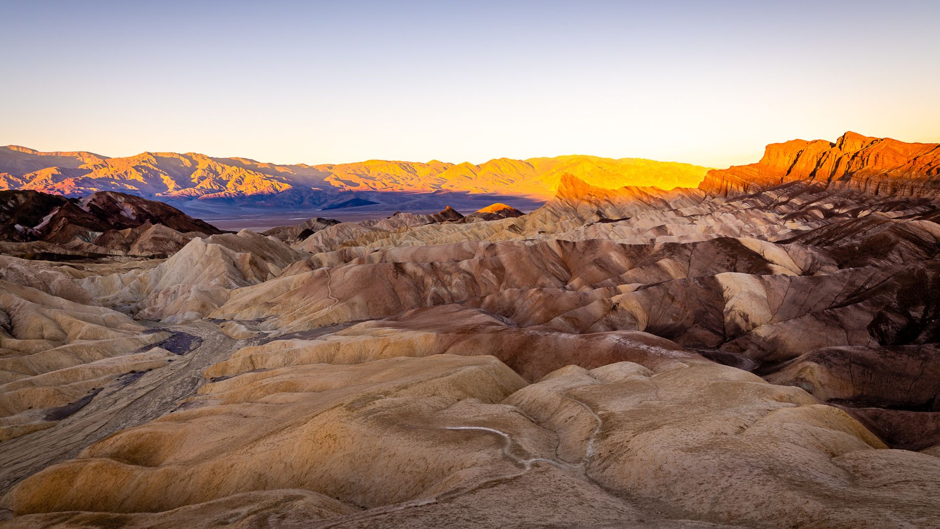 An expansive panoramic view of the rugged, eroded badlands of Death Valley National Park at sunrise or sunset, with warm golden light illuminating the peaks and valleys of the barren landscape and distant mountains under a clear sky.