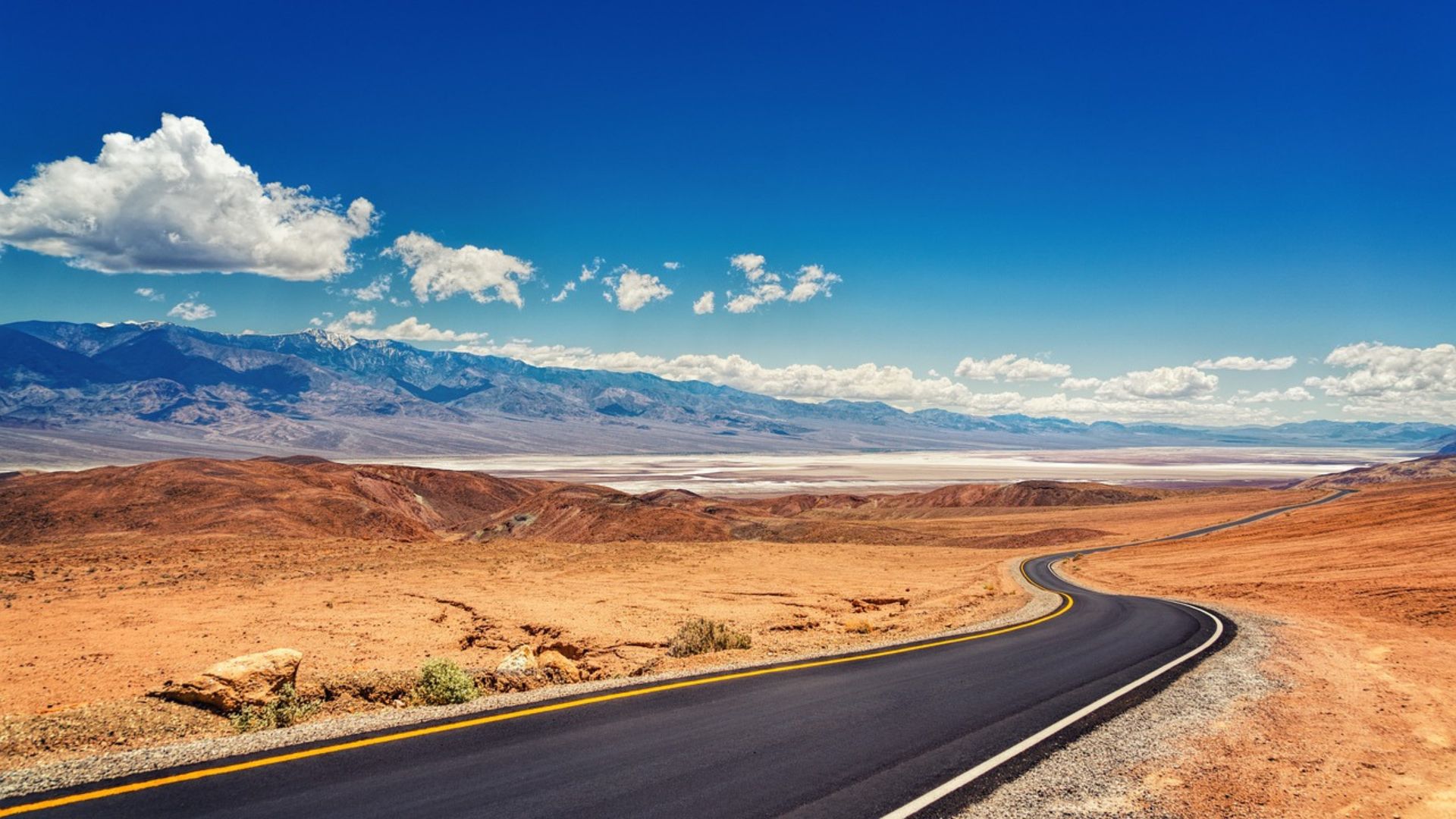 A winding asphalt road with a yellow center line and white edge lines curves through a vast, arid desert landscape under a clear blue sky with scattered white clouds. In the distance, rugged, light-colored mountains rise, and the ground appears to be a salt flat or dry lakebed leading up to their base. The foreground features reddish-brown, rocky desert terrain.