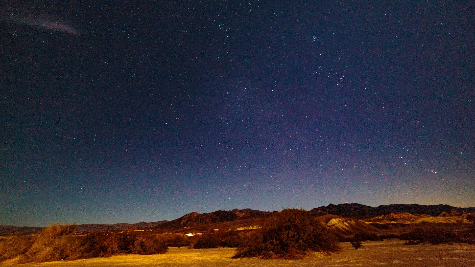 A wide-angle shot of a dark night sky filled with countless stars and a faint comet or meteor trail, above a desert landscape with low-lying brush and distant mountains silhouetted against the horizon. The foreground is dimly lit, revealing the texture of the desert floor.