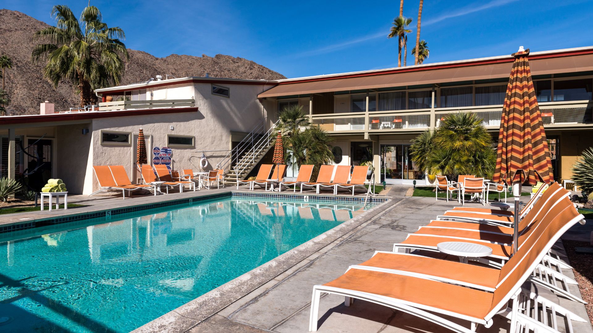 A vibrant outdoor scene at the Del Marcos Hotel in Palm Springs, California, featuring a sparkling blue swimming pool surrounded by orange lounge chairs and umbrellas, with a mid-century modern hotel building and palm trees under a clear sky, set against the backdrop of mountains.