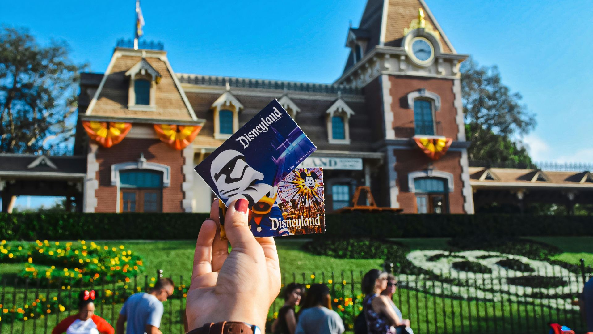 Hand holding Disneyland tickets with Star Wars and Donald Duck designs in front of the Disneyland Park entrance, featuring the Main Street, U.S.A. Train Station and Mickey floral display during daytime.