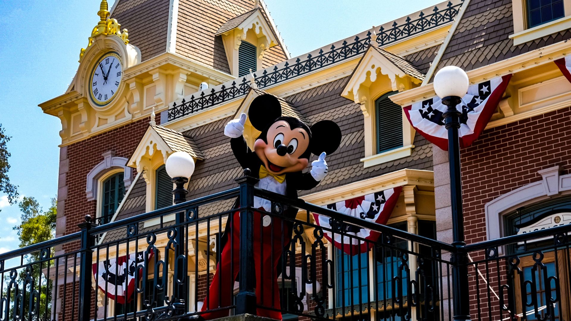 Mickey Mouse character in a tuxedo on a balcony at Disneyland Park in Anaheim, California, with the Main Street, U.S.A. train station building and clock tower in the background, ideal for Disney travel and theme park content.