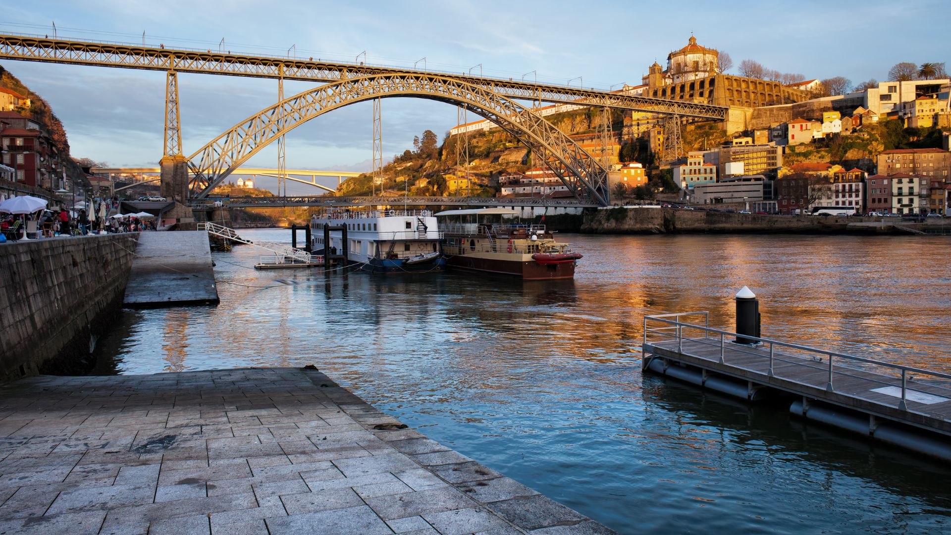 A wide shot of the iconic Dom Luís I Bridge spanning the Douro River in Porto, Portugal, connecting Porto to Vila Nova de Gaia, with boats docked along the riverbank and historic buildings visible on the hillsides under a partly cloudy sky.