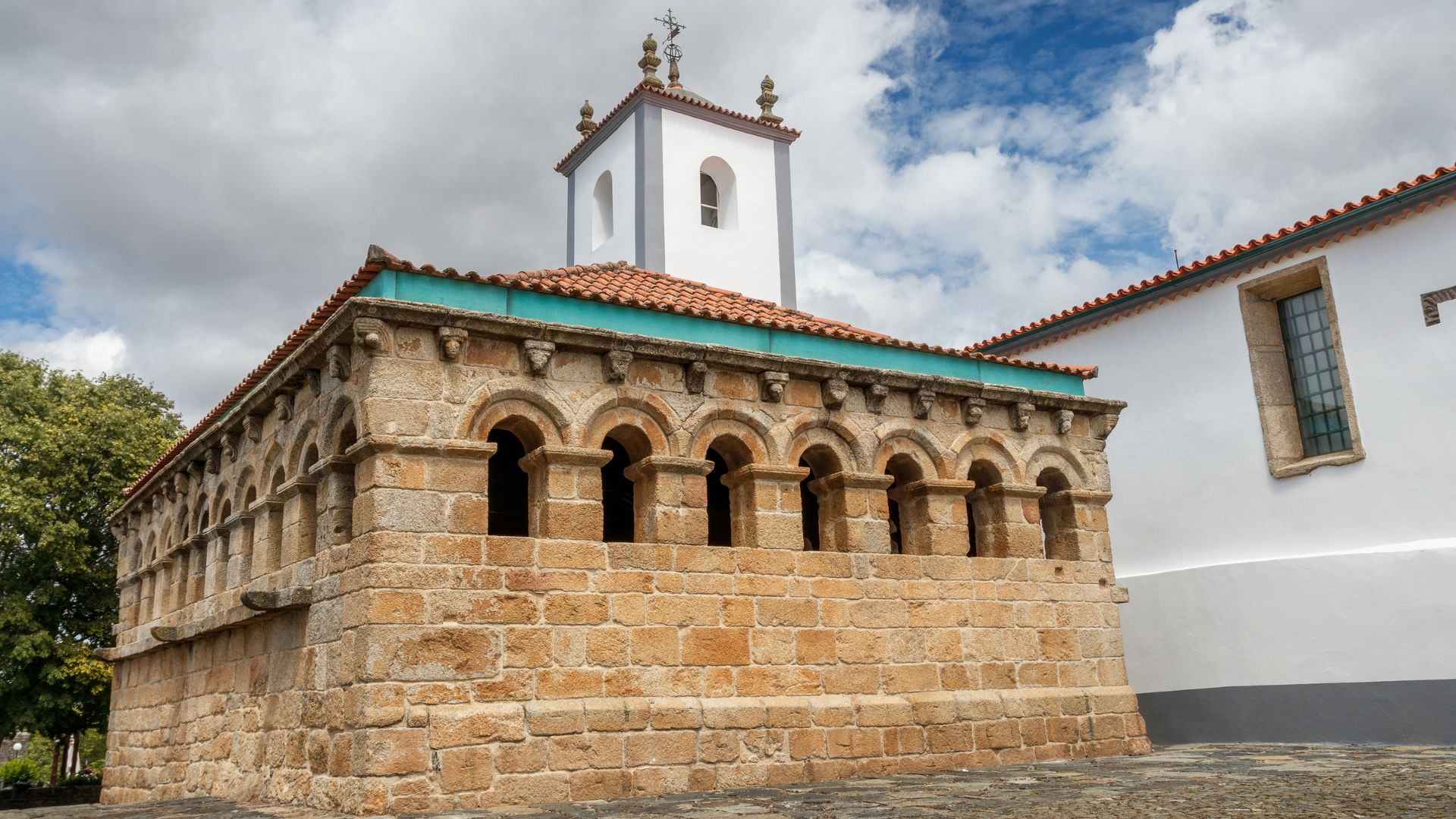 A low-angle shot of the Domus Municipalis in Bragança, Portugal, a Romanesque stone building with arched windows on its upper level and a white bell tower rising behind its red-tiled roof, set against a partly cloudy sky.