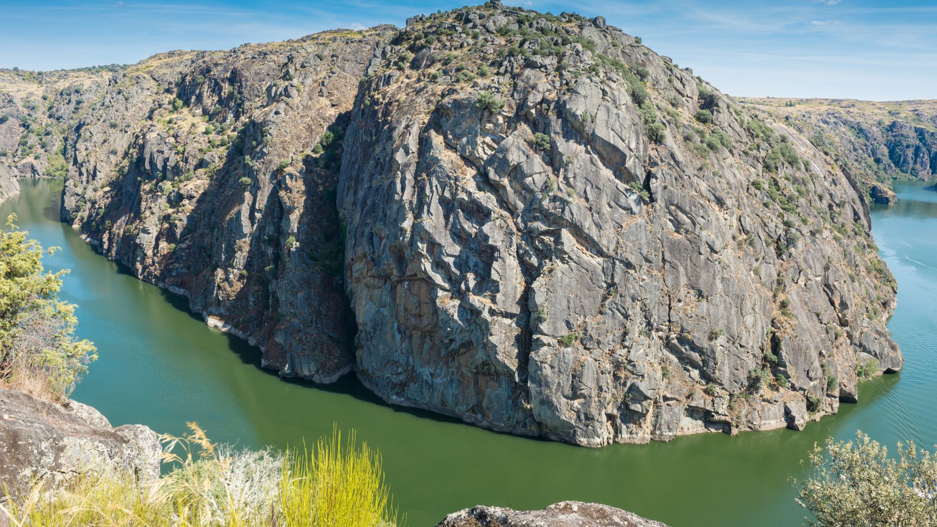 A wide shot of a river winding through a deep, rocky gorge with steep, tree-covered cliffs under a clear blue sky.