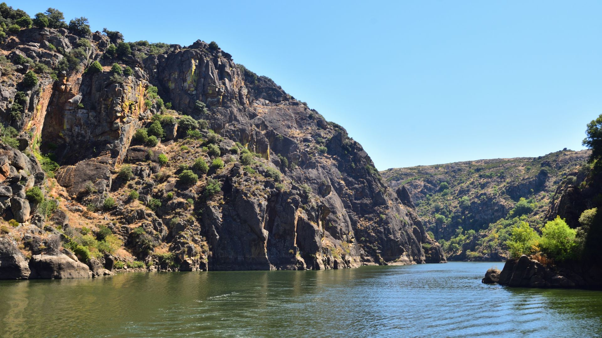 A wide shot of a river winding through a deep, rocky gorge with steep, tree-covered cliffs under a clear blue sky.