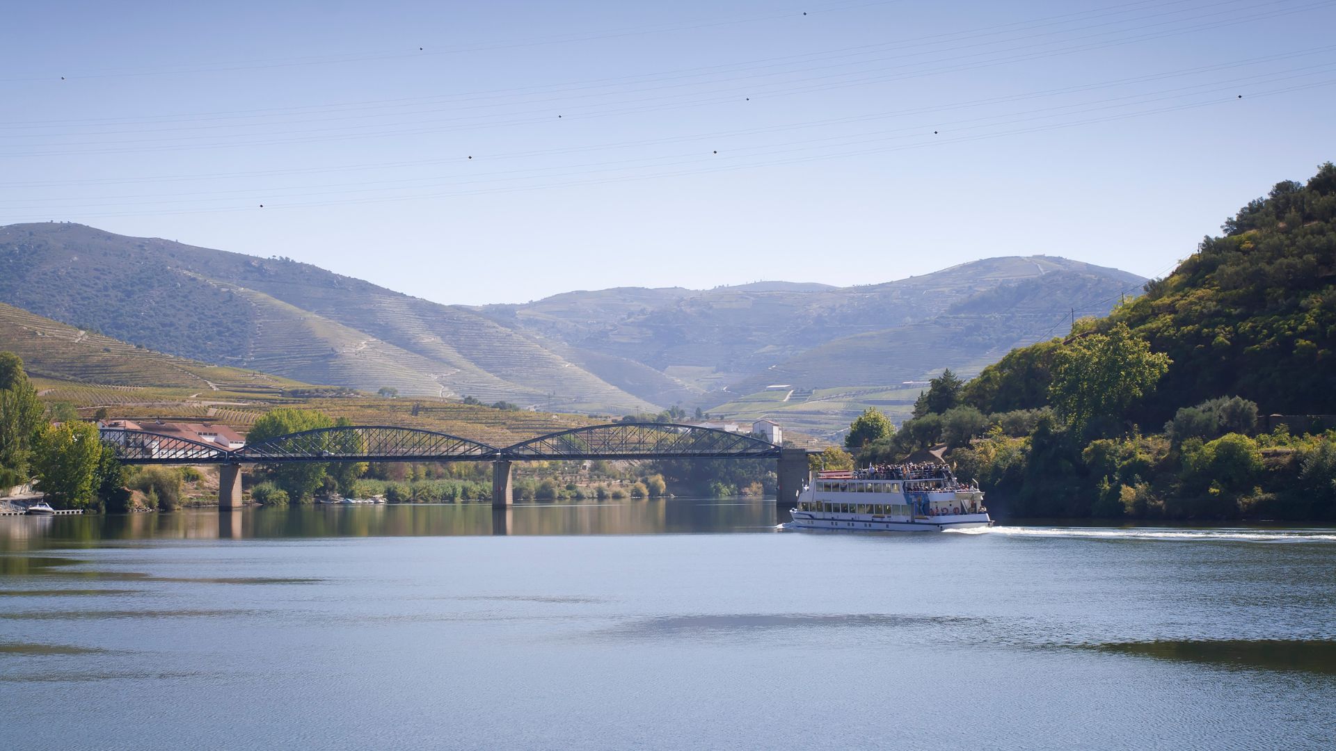 A scenic view of the Douro River in Portugal, featuring a bridge, a tour boat, and terraced vineyards on the surrounding hills under a clear sky. 