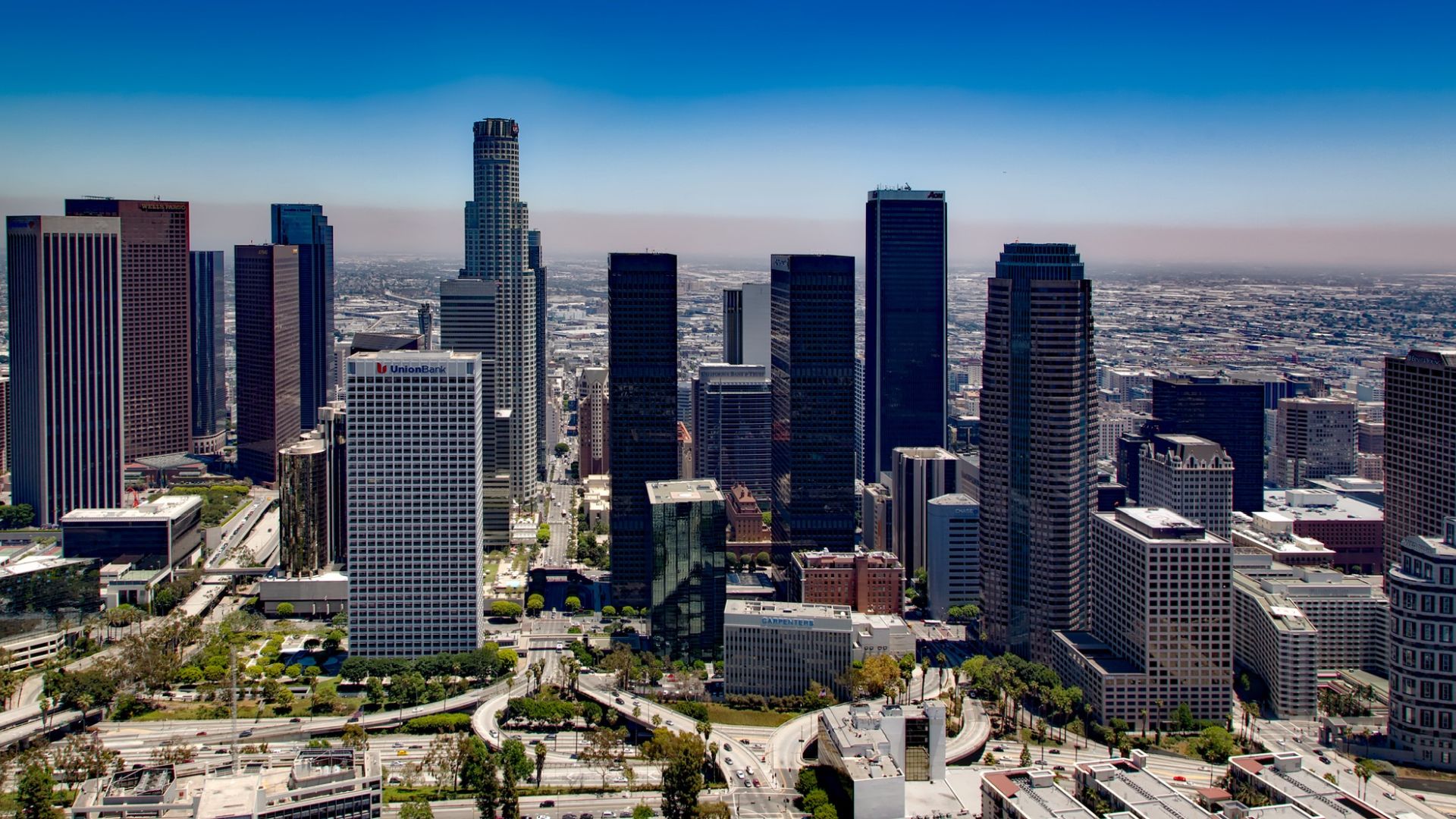 An aerial view of the Downtown Los Angeles skyline under a clear blue sky, featuring numerous skyscrapers including the prominent U.S. Bank Tower and other high-rise buildings, with freeways and urban infrastructure visible in the foreground. 