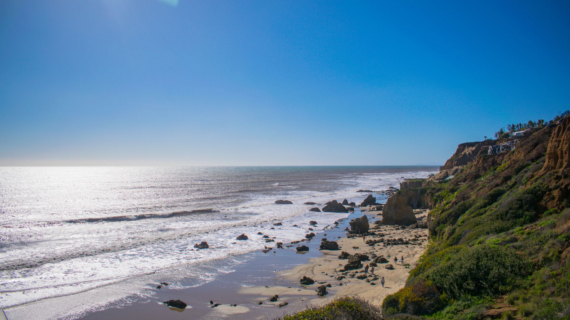 A sunny, wide-angle shot of El Matador State Beach in Malibu, California, showing the Pacific Ocean with waves crashing onto the sandy shore dotted with large rock formations and tide pools, alongside a steep, vegetated cliff face.