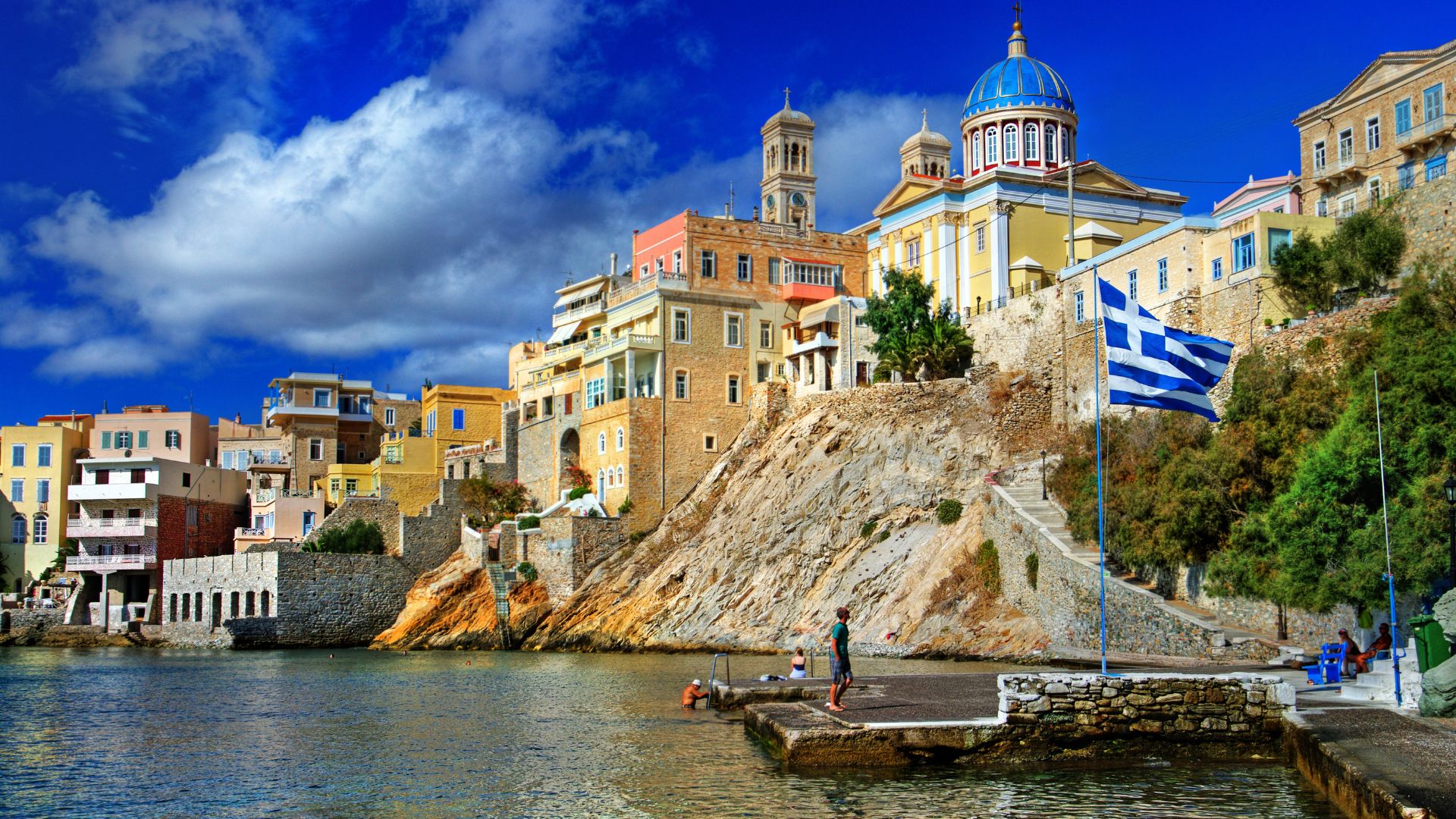 Ermoupoli, Syros Island, Greece: A colorful coastal town with a Greek flag and a blue-domed church.