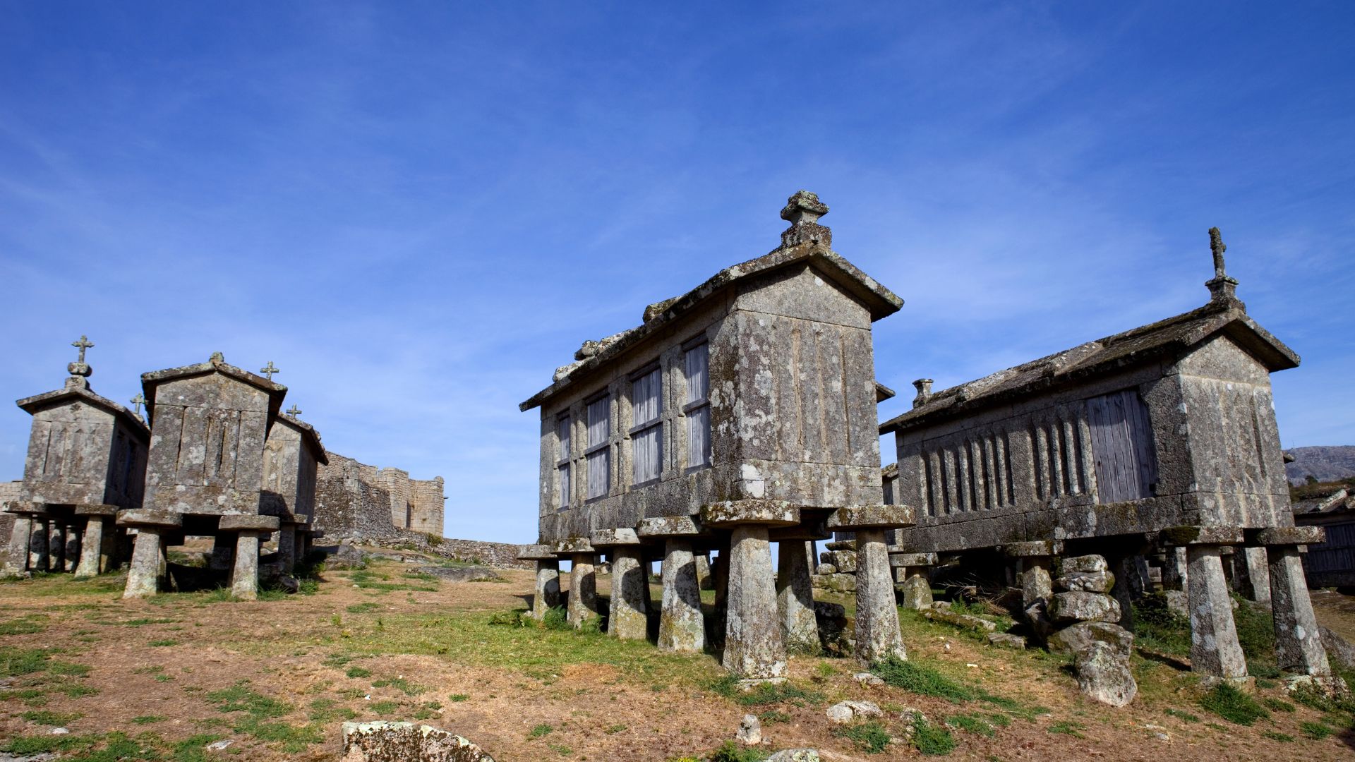 A group of ancient stone granaries, known as espigueiros, stand on elevated stone pillars against a clear blue sky in Lindoso, Portugal, with a distant castle ruin visible on a hill.