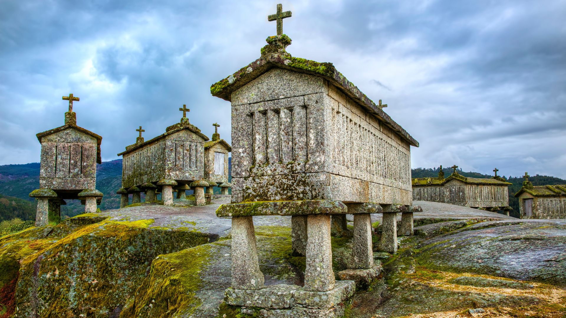 A panoramic view of the Espigueiros de Soajo, a collection of ancient stone granaries with cross-shaped finials on their roofs, standing on a rocky outcrop under a cloudy sky in the village of Soajo, Portugal.