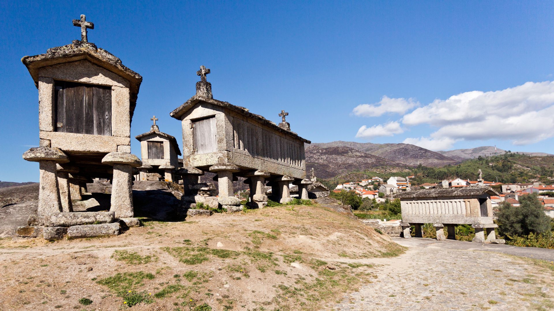 Ancient stone granaries (espigueiros) with crosses on top, set on a hillside in the village of Soajo, Portugal, with mountains and a distant village in the background under a blue sky.