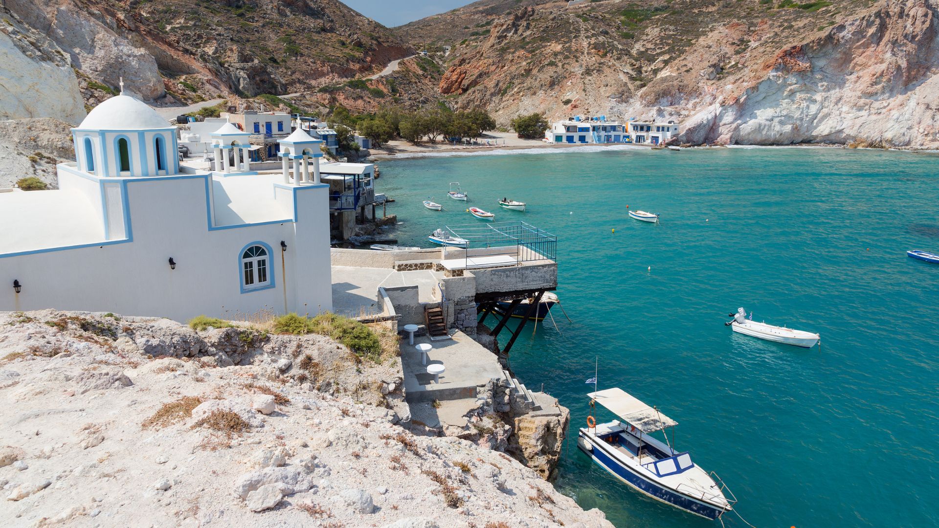 Scenic view of Firopotamos in Milos, Cyclades, Greecce featuring white building, mountains, blue waters, and boats.