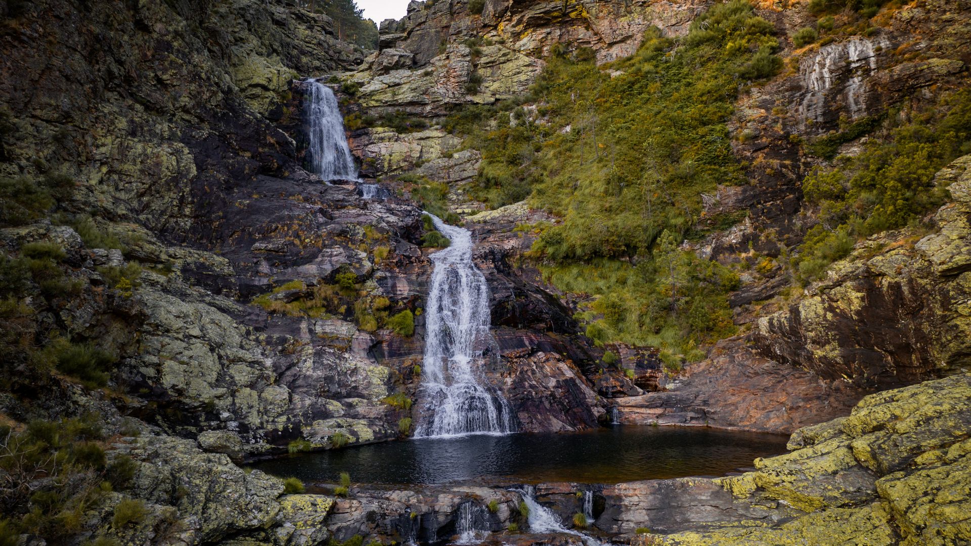 A multi-tiered waterfall cascades down a rocky, moss-covered cliff face into a dark pool at its base, surrounded by lush green and grey-toned natural rock formations.