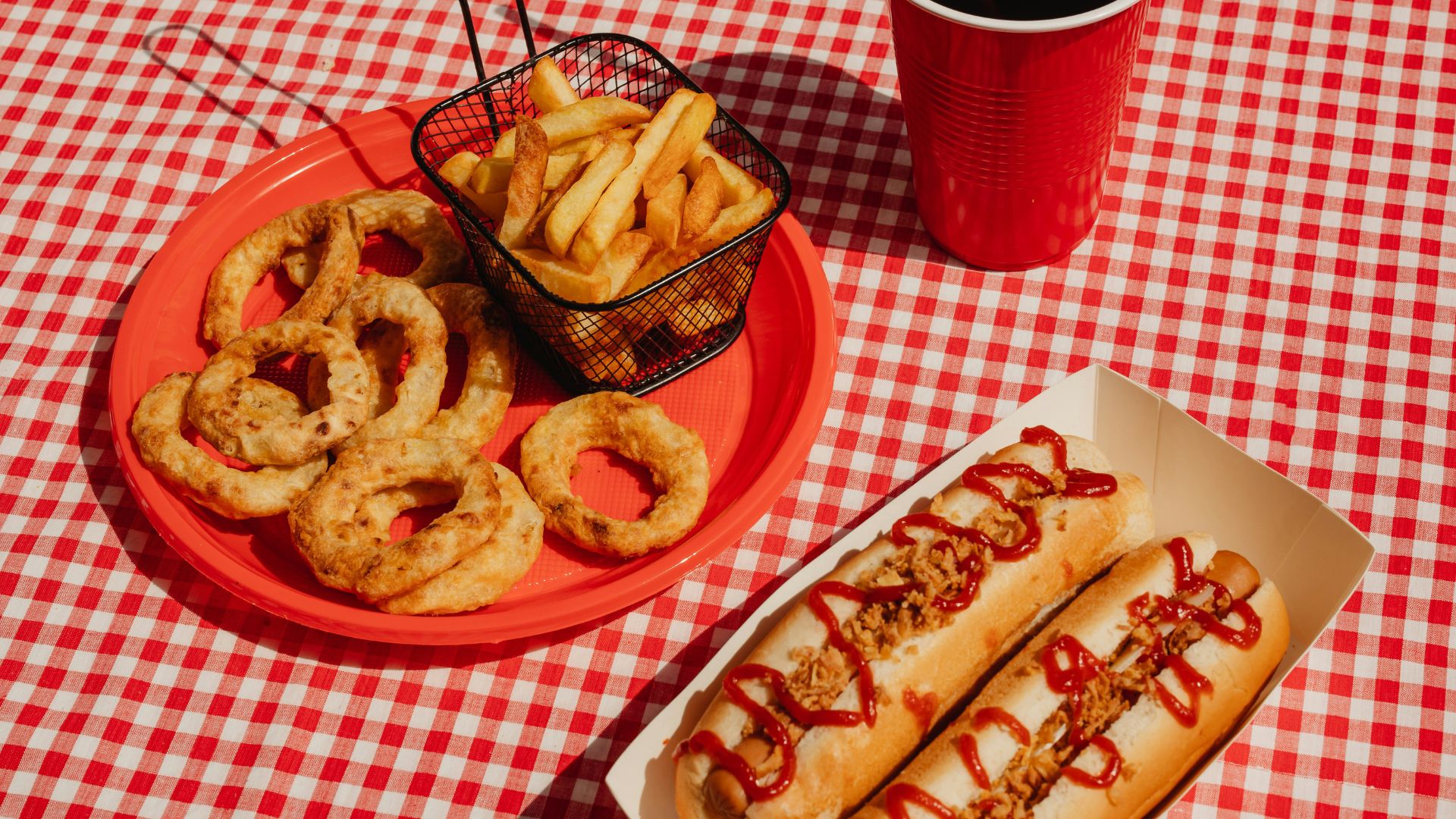 Picnic spread with hot dogs, crispy french fries in a wire basket, golden onion rings, and a refreshing soda on a classic red and white checkered tablecloth, perfect for outdoor dining or summer events.