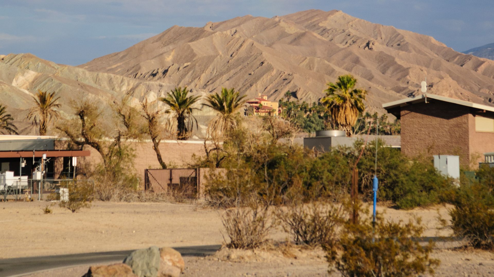 Deep blue sky over a desert landscape in Death Valley National Park. In the foreground, sparse desert vegetation and a few buildings are visible, with rugged, arid mountains rising in the background under the clear sky.
