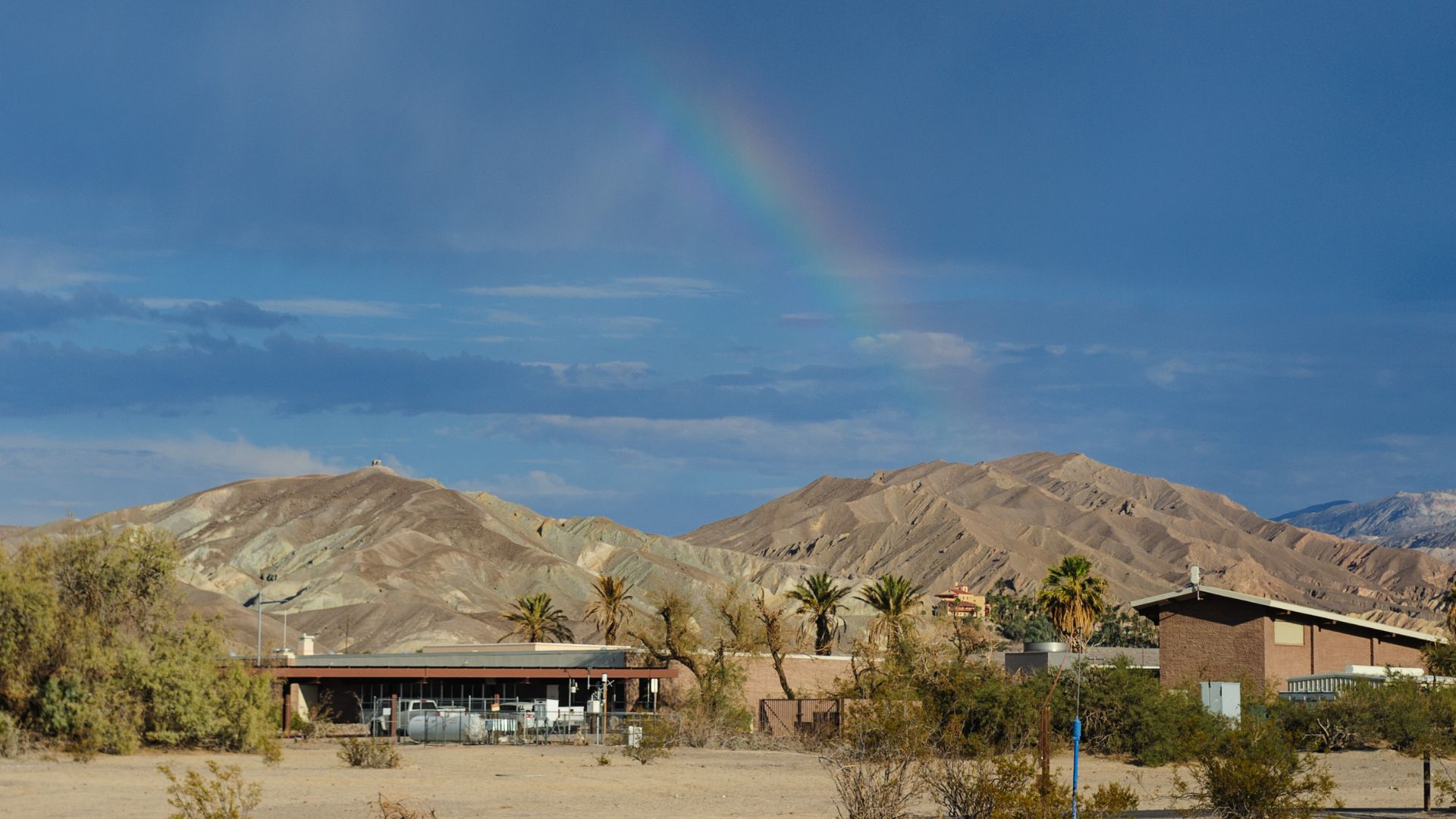 A vibrant rainbow arches across a deep blue sky over a desert landscape in Death Valley National Park. In the foreground, sparse desert vegetation and a few buildings are visible, with rugged, arid mountains rising in the background under the clear sky.