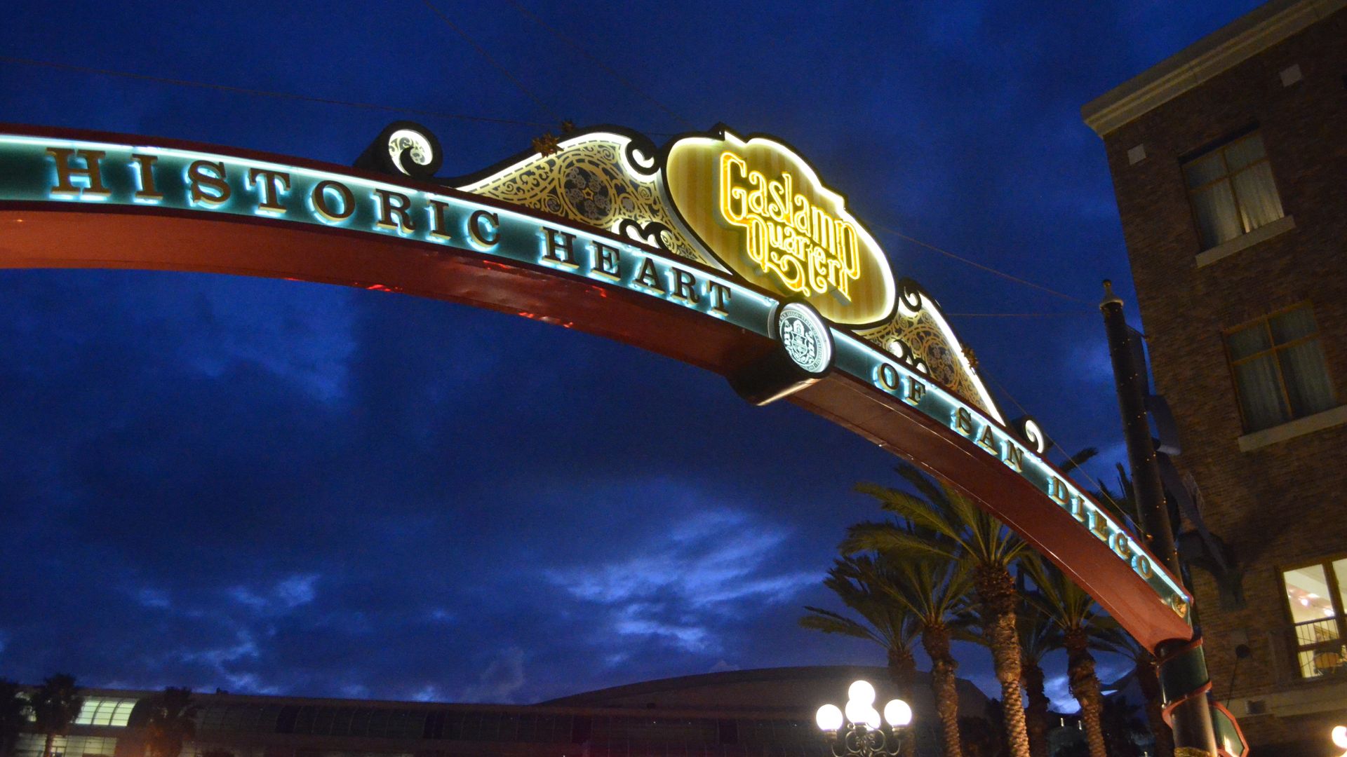 "Nighttime view of the illuminated 'Historic Heart of San Diego Gaslamp Quarter' archway sign, with palm trees and a building visible against a dark blue sky.