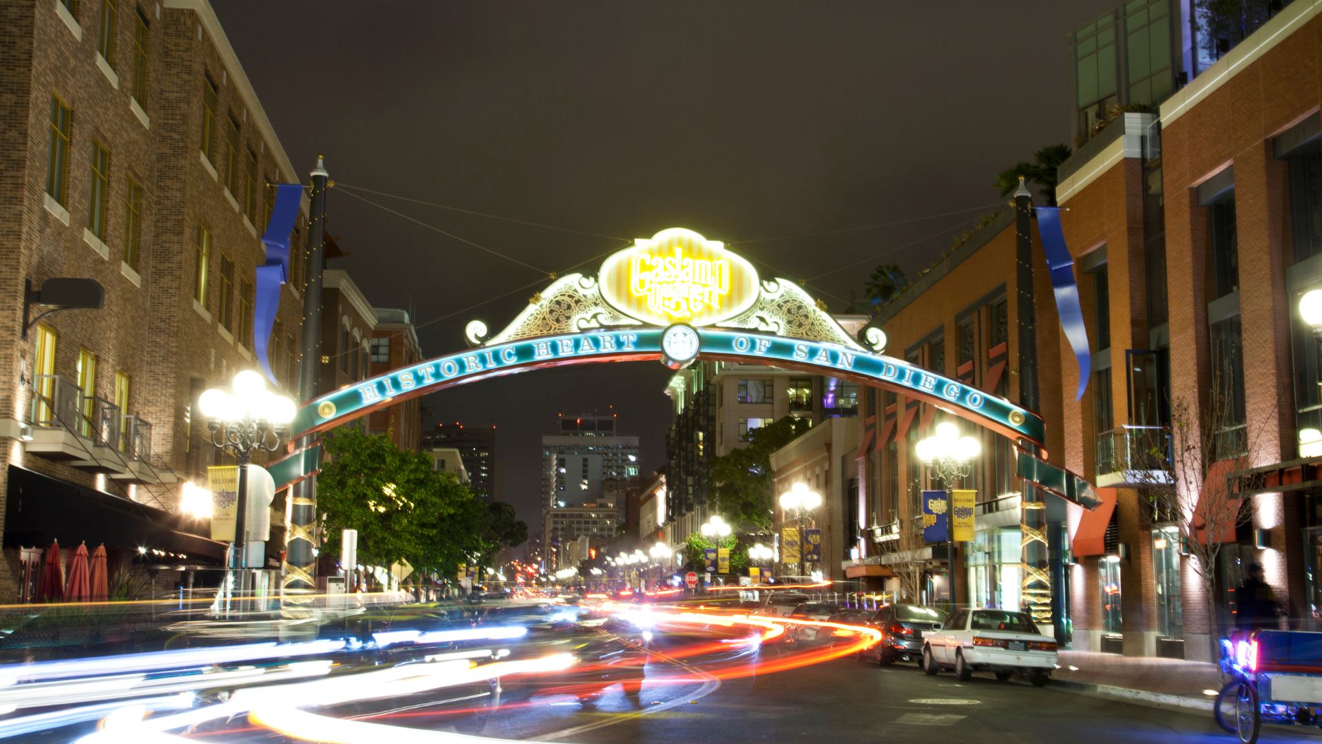 A vibrant night scene of the Gaslamp Quarter in San Diego, featuring the illuminated "Historic Heart of San Diego" archway sign spanning across a street filled with light trails from moving vehicles, flanked by historic buildings and streetlights. 