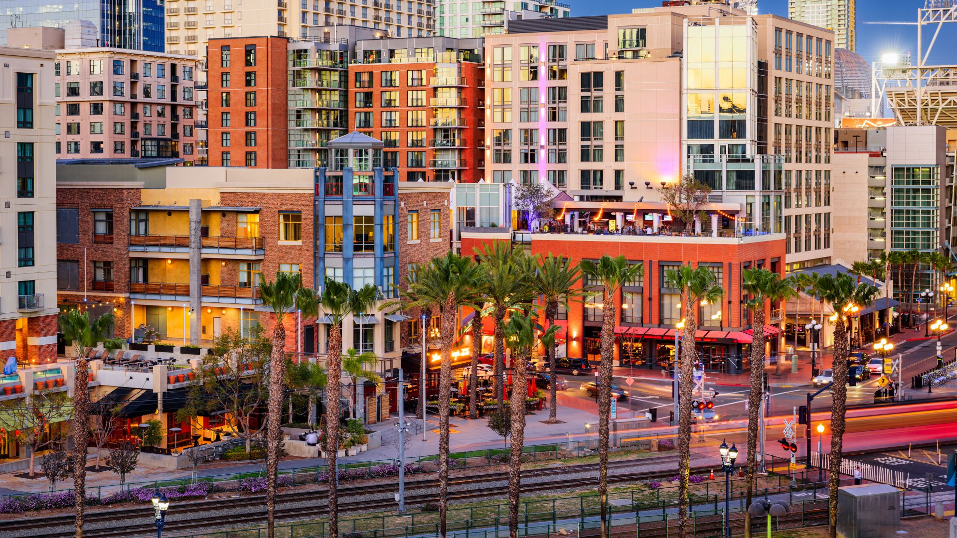 An elevated view of the Gaslamp Quarter in San Diego, California, showcasing multi-story buildings, palm trees, and streetlights along a bustling street at dusk or night, with light trails from moving vehicles and visible train tracks in the foreground.