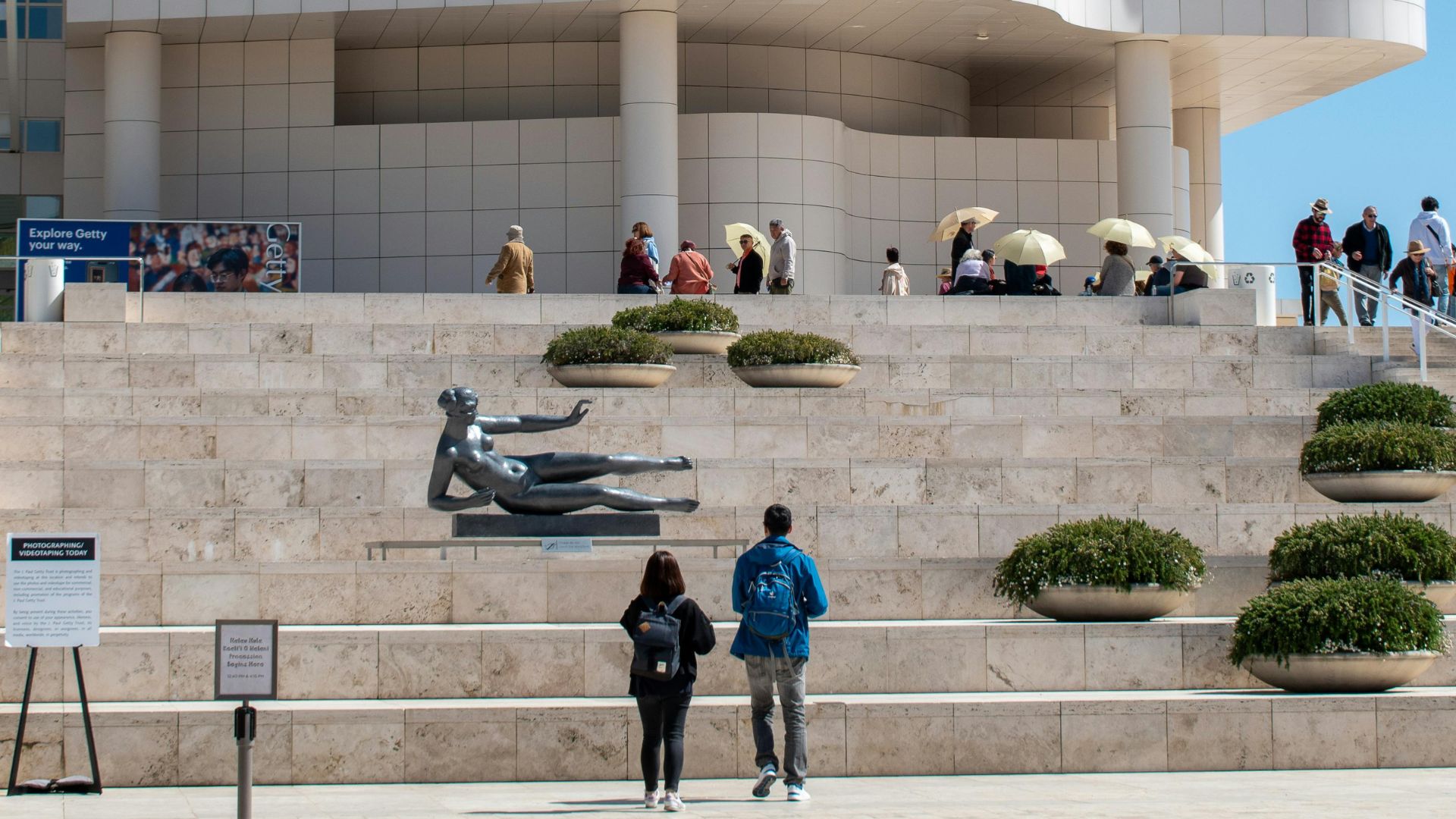 Exterior view of the Getty Center in Los Angeles, featuring its distinctive white modern architecture, a bronze sculpture of a reclining female figure ('Air' by Aristide Maillol), and visitors ascending the grand staircase under a clear sky