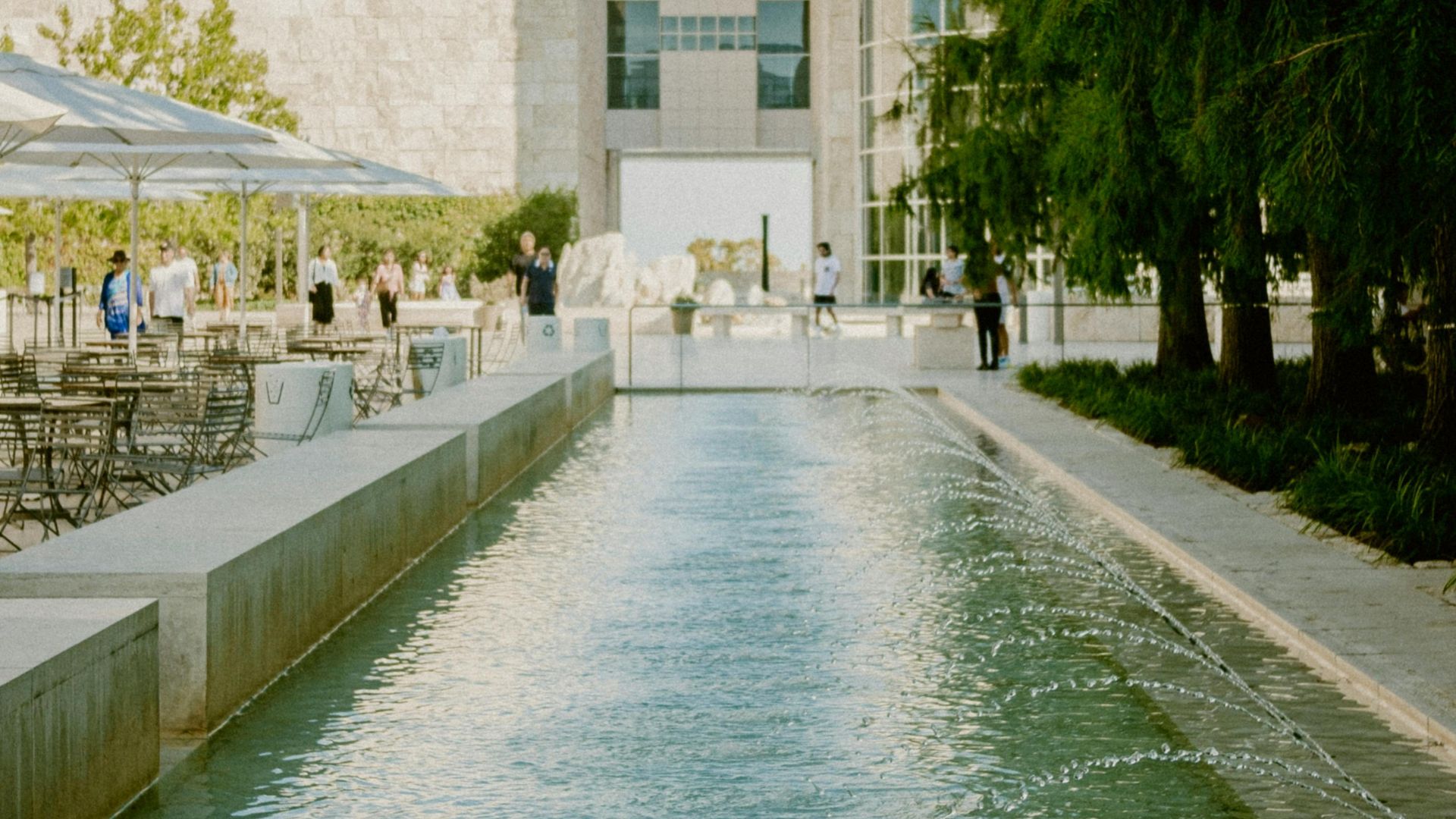 A serene outdoor shot of the Getty Center in Los Angeles, featuring a long, reflective water channel leading towards the modern, light-colored stone architecture of the museum buildings, with lush green trees lining the right side and outdoor seating with umbrellas on the left.