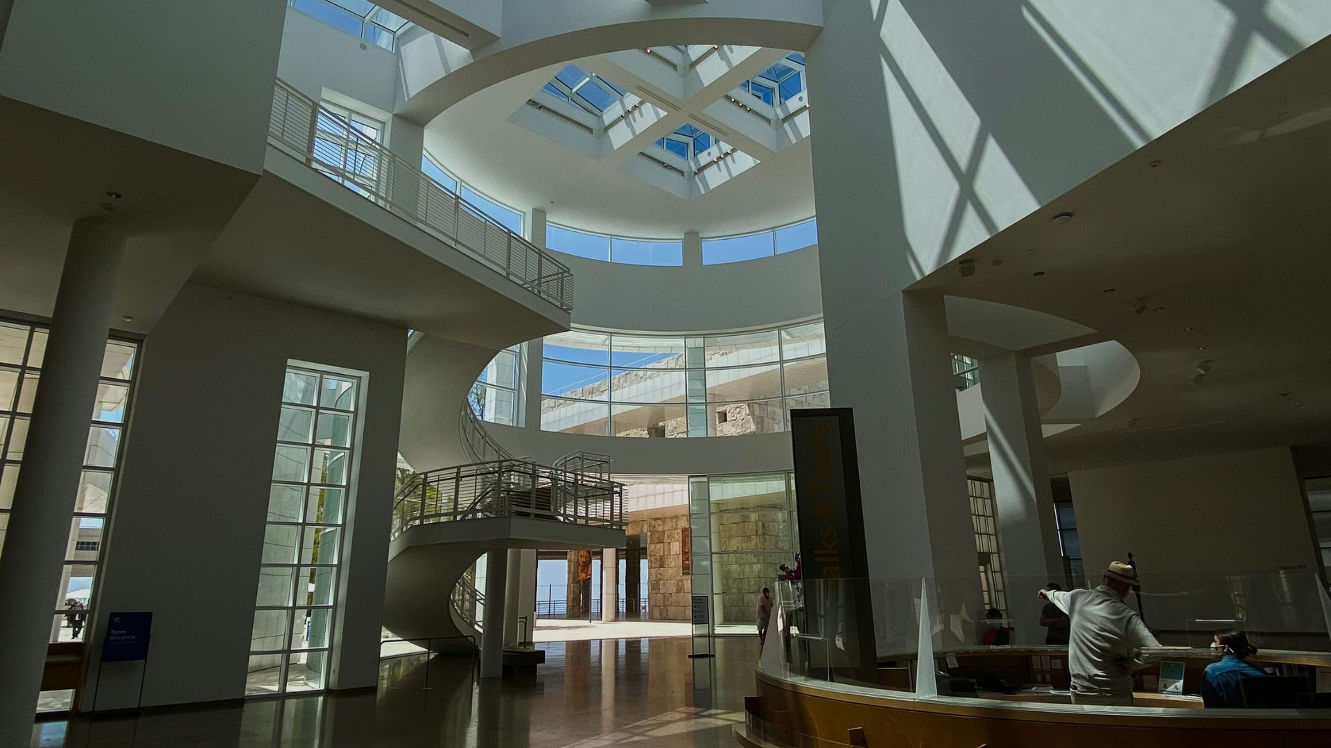 Grand entrance hall of the Getty Center Museum in Los Angeles, featuring a high glass ceiling, modern white architecture, spiral staircase, and a wooden information desk with staff.