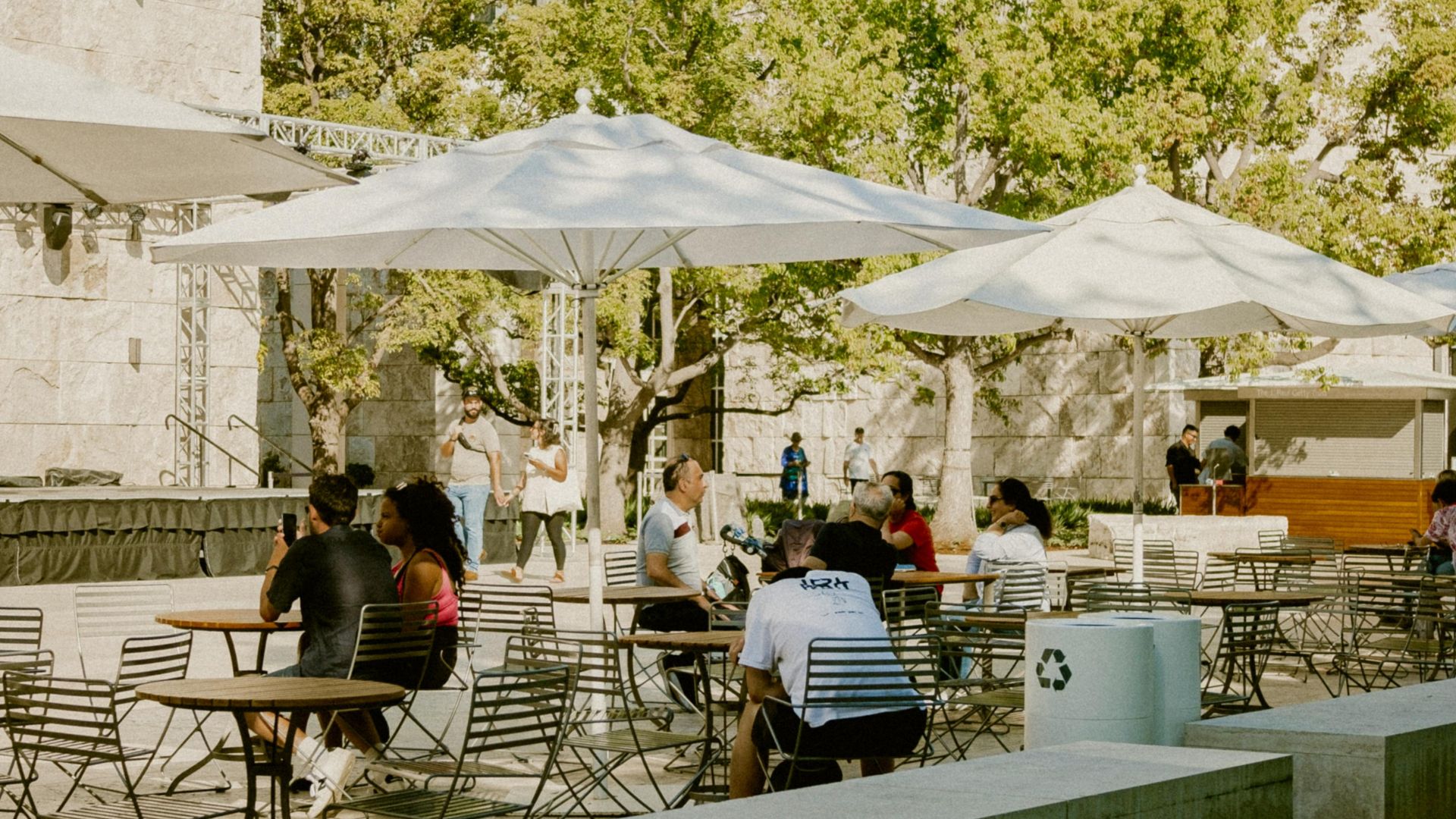 Outdoor dining area at the Getty Center in Los Angeles, featuring white umbrellas, tables with chairs, and people enjoying a sunny day by a reflective pool with modern stone architecture and lush green trees in the background.