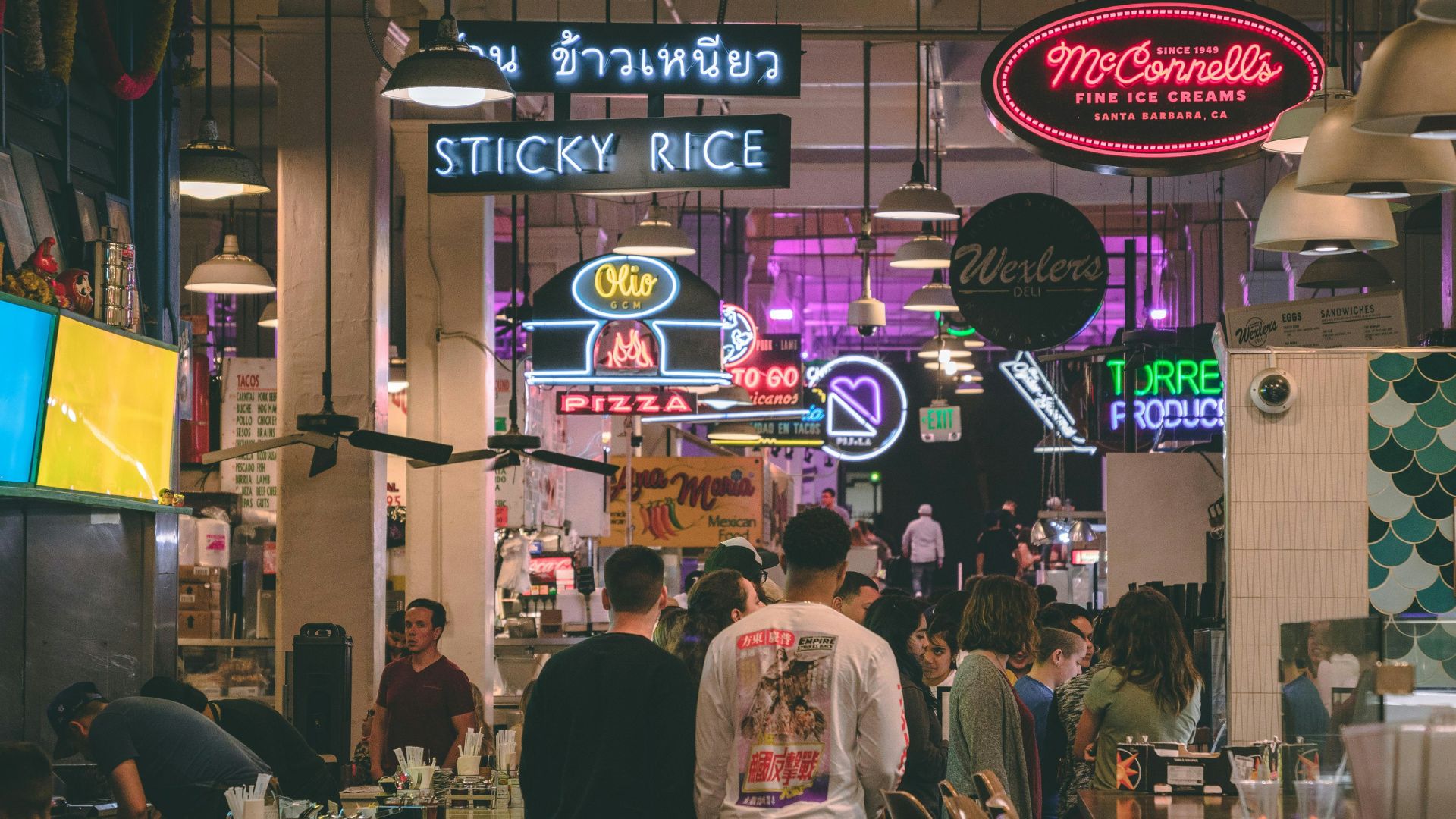 A vibrant, bustling indoor market scene with various food stalls and neon signs, including "STICKY RICE," "Olio PIZZA," "McConnell's FINE ICE CREAMS," and "Wexler's," with people gathered at counters and walking through the aisles.