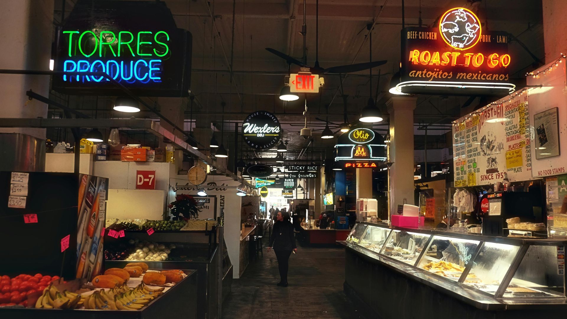 A bustling indoor market with various food stalls and vendors under neon signs, including "Torres Produce" and "Roast To Go," featuring display cases of food and a person walking down an aisle.