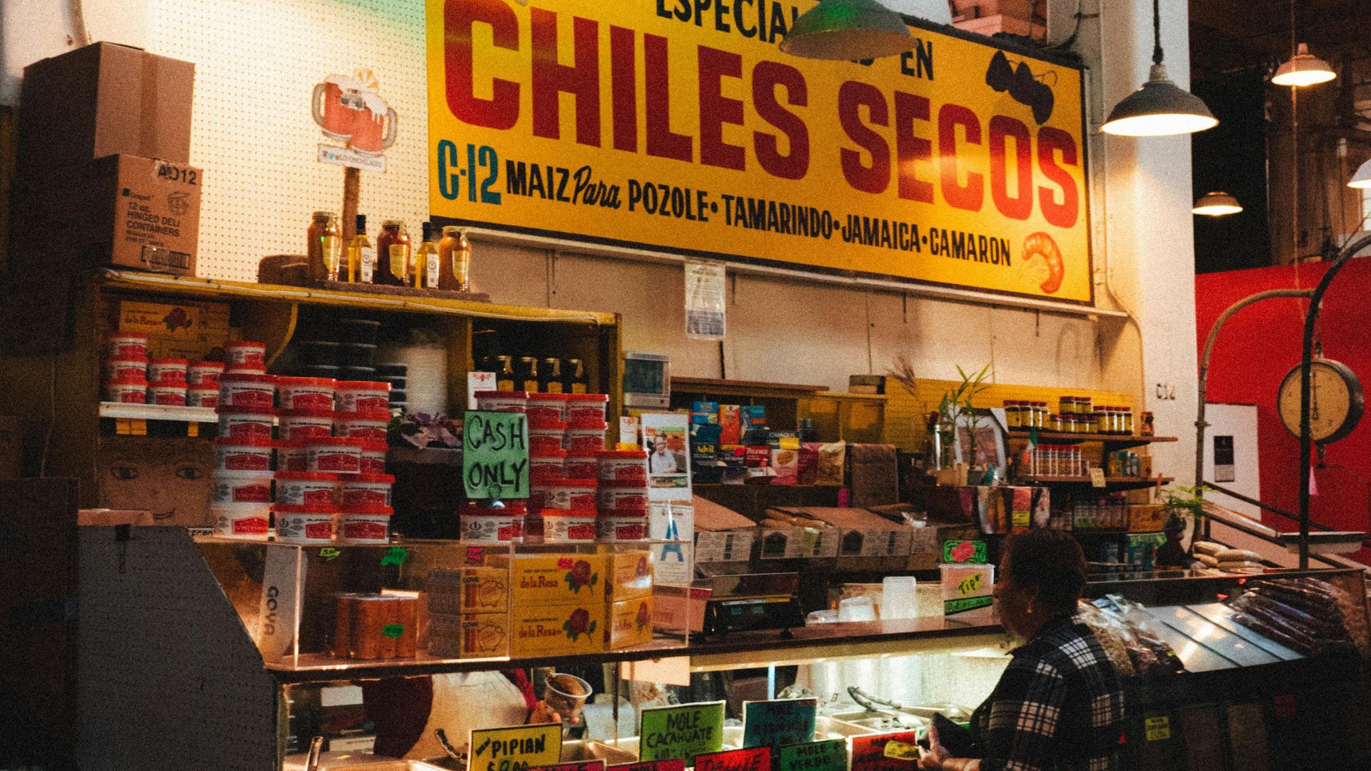 A vibrant indoor market stall with shelves filled with various packaged goods, primarily dried chiles and mole pastes. Above the stall, a neon sign reads "MOLES" and "CHILES SECOS." Below it, a large yellow banner reiterates "CHILES SECOS" in red lettering. A person is visible on the right side of the image, near the counter.