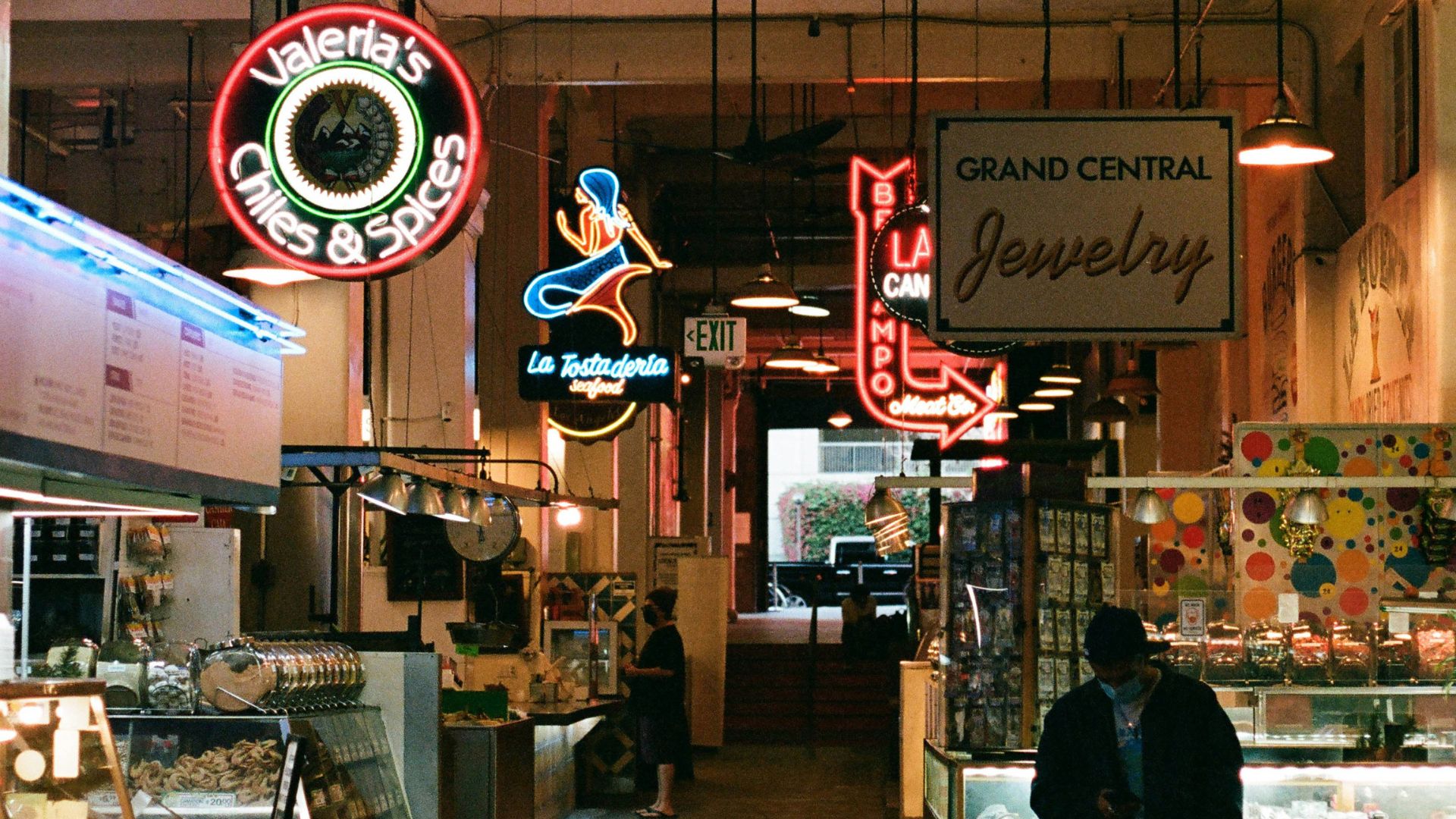 Indoor shot of Grand Central Market in Los Angeles, featuring various food stalls and neon signs like 'Valeria's Chiles & Spices' and 'Grand Central Jewelry,' with a person standing near a display case and another walking in the background.