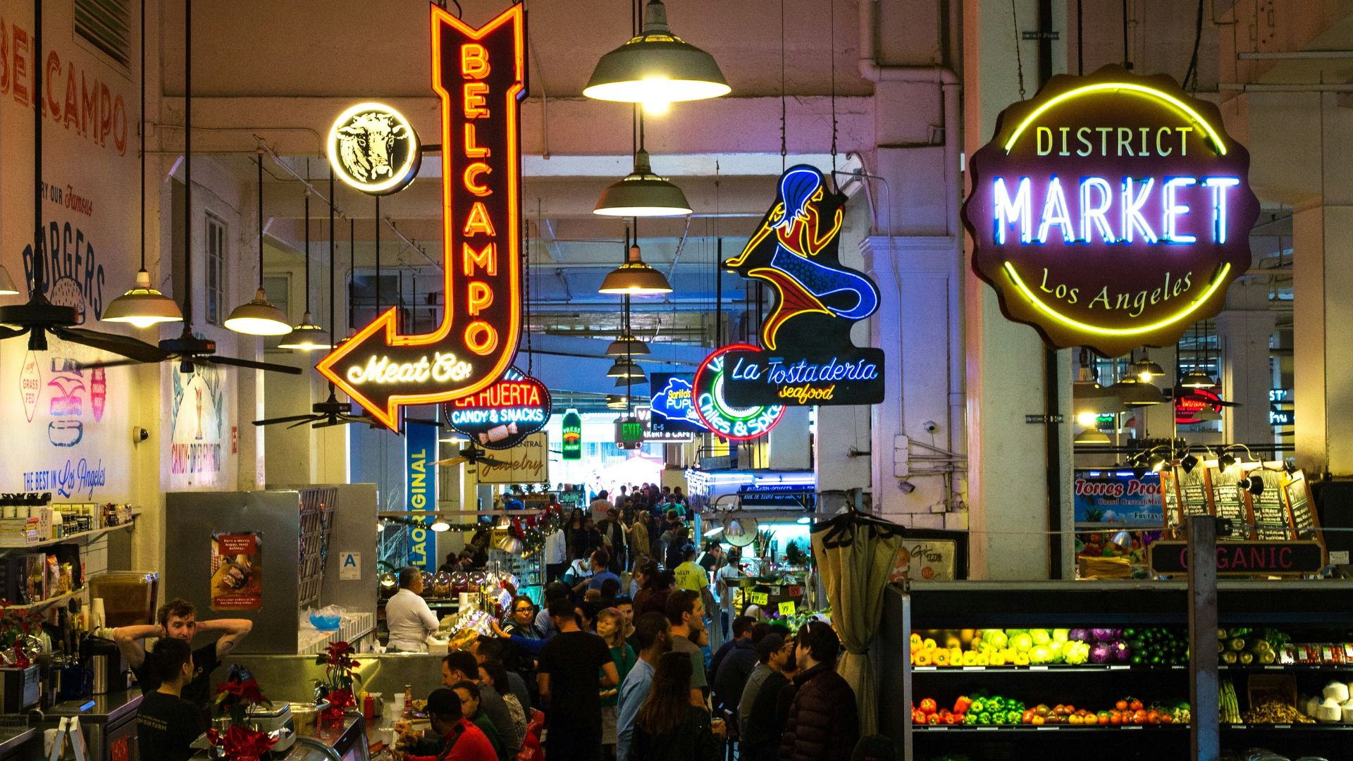 Vibrant interior of Grand Central Market in downtown Los Angeles, featuring diverse food stalls, neon signs like "BELCAMPO" and "DISTRICT MARKET Los Angeles," and a bustling crowd of shoppers and diners enjoying the historic food hall atmosphere.